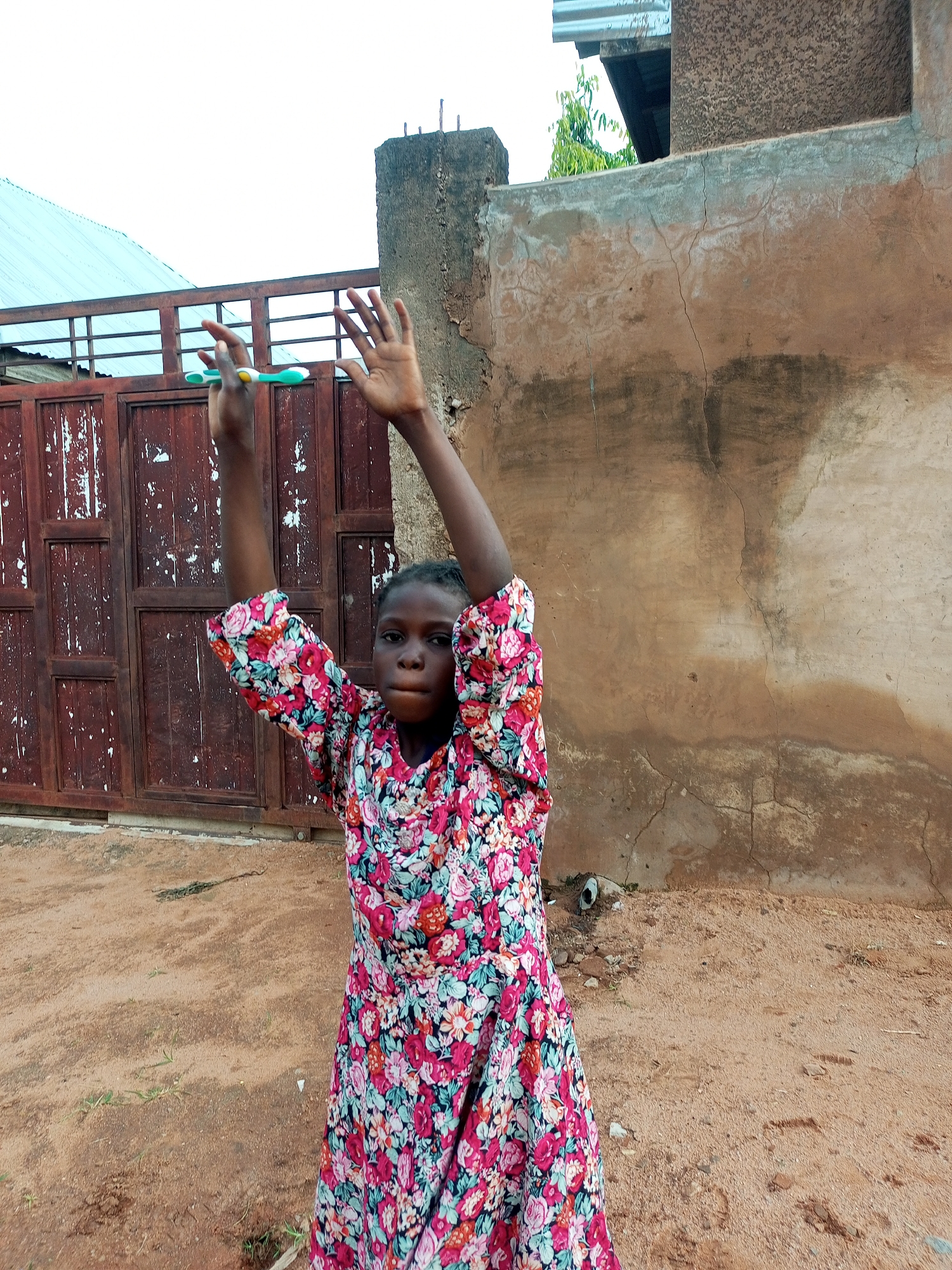 A young Black girl wearing a floral print dress stands outside a building in Tempure, Nigeria. She has her arms raised and appears to be holding a toothbrush in her left hand. The building has a textured, brown facade and a weathered, red metal gate behind the girl. The ground is covered in dirt and sand.