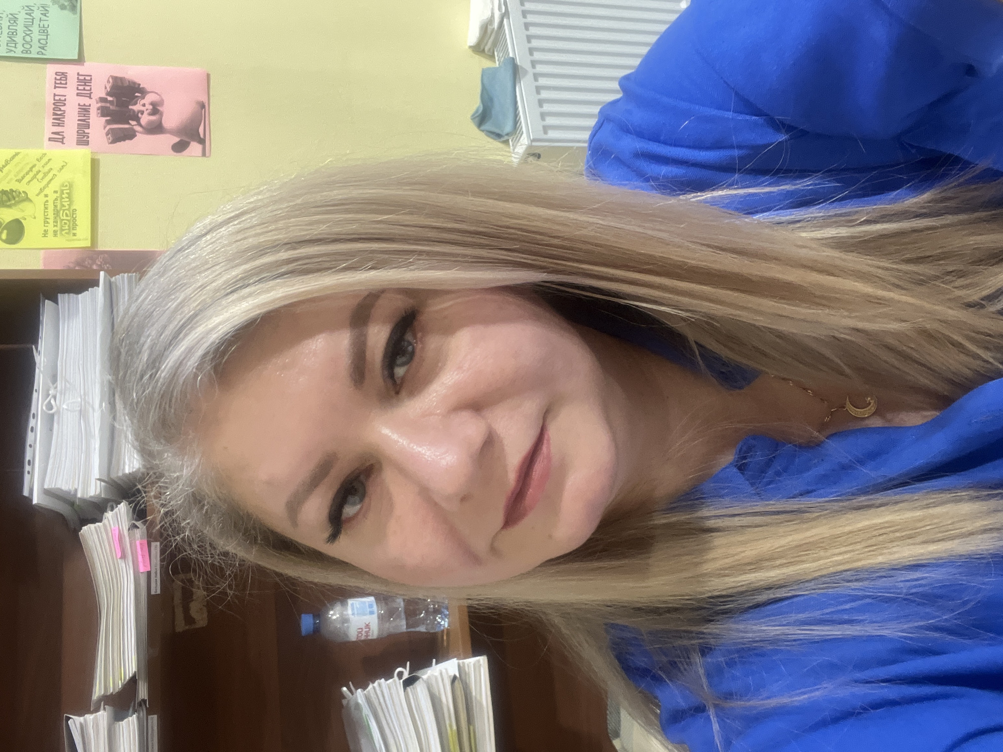 Here is a detailed description of the image:
The image is a selfie of a woman with blonde hair wearing a blue top. She has light skin, grey eyes, and is wearing makeup, including eyeliner and lipstick. She is smiling slightly.
In the background, there is a wooden shelf stacked with papers. A bottle of water with Cyrillic writing on the label is visible on the shelf. On the wall behind the shelf, there are several small posters with Cyrillic text and images. One poster has a cartoon cat with stacks of money and the text "ДА НАКРОЕТ ТЕБЯ ШУРШАНИЕ ДЕНЕГ" (May the rustle of money cover you).
Based on the Cyrillic text and general appearance, the location is likely in Russia. Without additional details, it's impossible to definitively determine if it is Izhevsk.