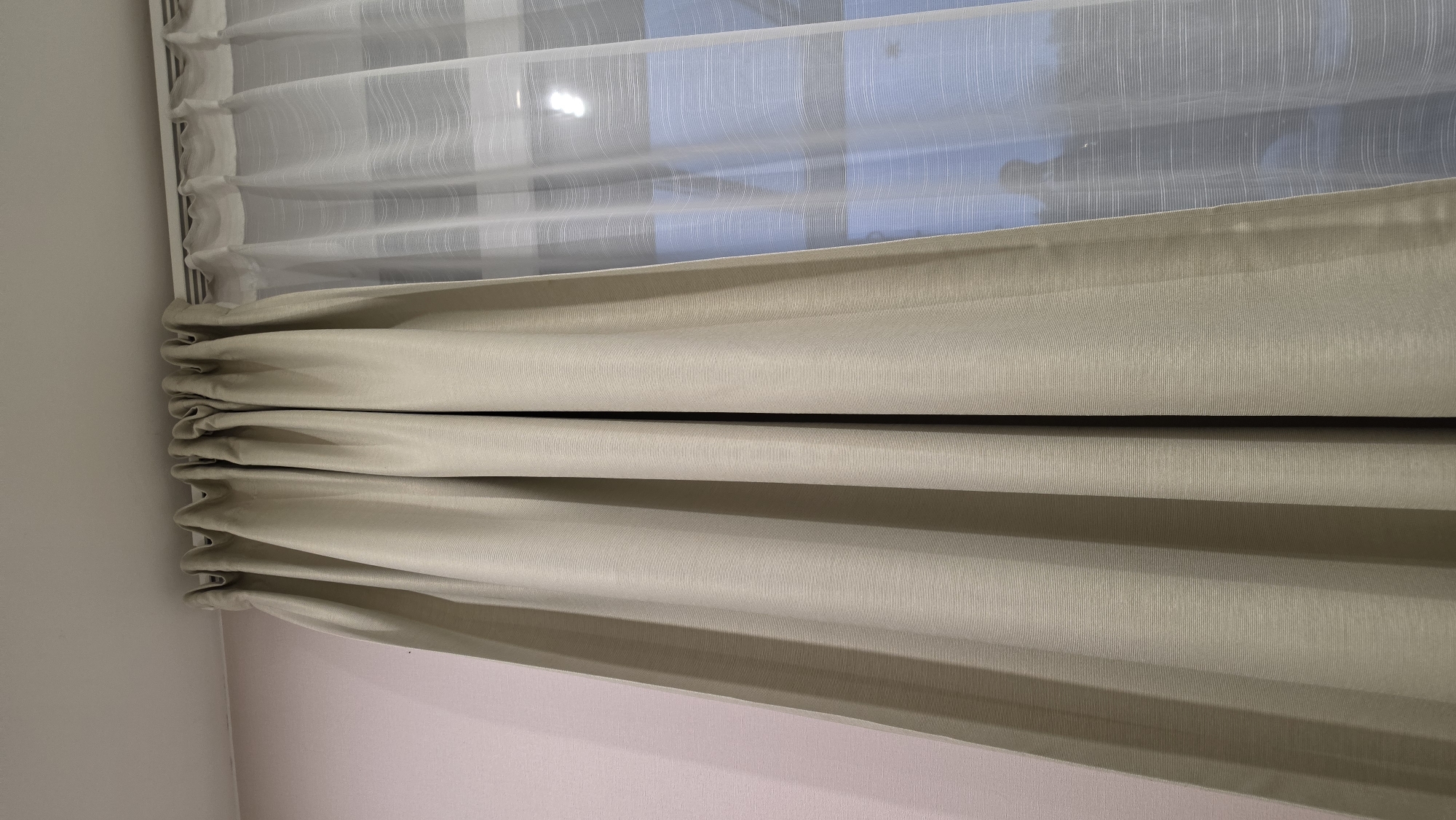 This image captures an interior view of a window, likely in a room located in Ihringshausen, Germany. The main subjects are two layers of curtains and the surrounding wall.
The window features a double-layered curtain system. The inner layer, closest to the window, is a sheer, translucent white fabric with a fine, subtle vertical striped texture. Through this sheer curtain, a soft blue sky is visible, along with indistinct, darker shapes that suggest an outdoor view of objects like buildings or trees.
The outer layer is a thicker, opaque curtain in a light beige or khaki color. This curtain is partially drawn upwards, creating prominent horizontal folds, indicative of a Roman blind style or similar gathering mechanism. It also exhibits a subtle vertical ribbed or striped texture, mirroring the pattern of the sheer curtain. Both curtain layers are attached to a white window frame or curtain rail along the top and left side of the composition.
To the immediate left of the curtains, a plain white wall is visible. Below the lower edge of the beige curtain, a wall painted in a soft, light pink hue is shown.
The lighting suggests it is daytime, with a diffused light quality implying either an overcast day or an indirect light source. There are no people, explicit activities, specific weather details beyond the sky color, or visible text present in the image.