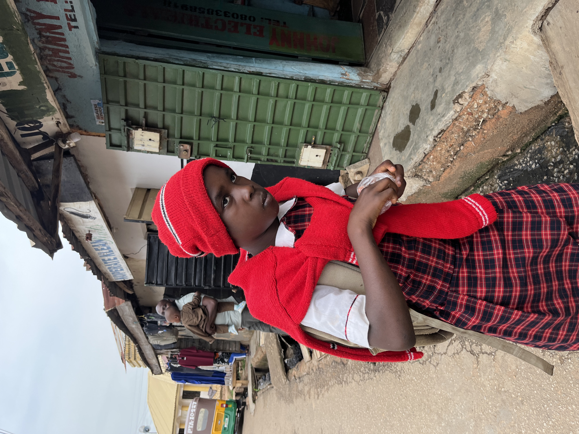 A medium shot, rotated 90 degrees clockwise, captures a street scene in what appears to be an urban or semi-urban market area under an overcast sky.
The primary subject is a young person, likely a child or pre-teen, positioned in the foreground. They wear a red knit beanie with white stripes, a red knitted sweater draped over their shoulders, a white shirt with red trim, and a red and black plaid skirt or dress. A beige backpack is visible on their back. They are holding a clear plastic bag in their right hand and look slightly upwards and to the right with a neutral, observant expression.
In the background, behind the young person, are several market stalls or shops. One prominent shop features a green corrugated metal door/shutter and a light blue painted facade above it. Another shop to the left has a more rudimentary structure with corrugated metal roofing. An adult male is partially visible in the background, standing or sitting near some hanging clothes, holding a brown jacket. Further back and to the right, a yellow and green vehicle is partially visible. The ground is a mix of concrete and dirt, with some debris and what appear to be small puddles.
Visible text includes: "JOHNNY ELECTR" and "TEL: 0803305891" on the blue shop facade. A white sign partially reads "SUPREME CLEAN". A sticker on the green door is difficult to decipher but appears to say "STRESS SERVICE" or similar. On the yellow vehicle, "PTA-10427" is visible. The scene depicts everyday activity in a bustling street environment.