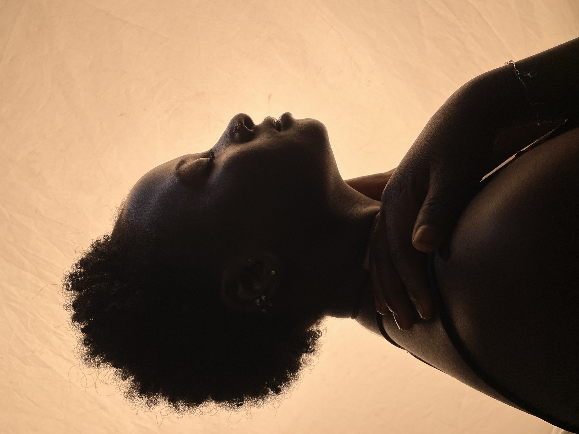 The image depicts a low-angle view of a Black woman with curly hair, lying down. Her head is tilted back, and her eyes are closed. Her arms are crossed over her chest. She has an earring or earrings visible. The background is a plain, light-colored surface. The location in the city of Jos, Nigeria is unknown and not inferable from the image.