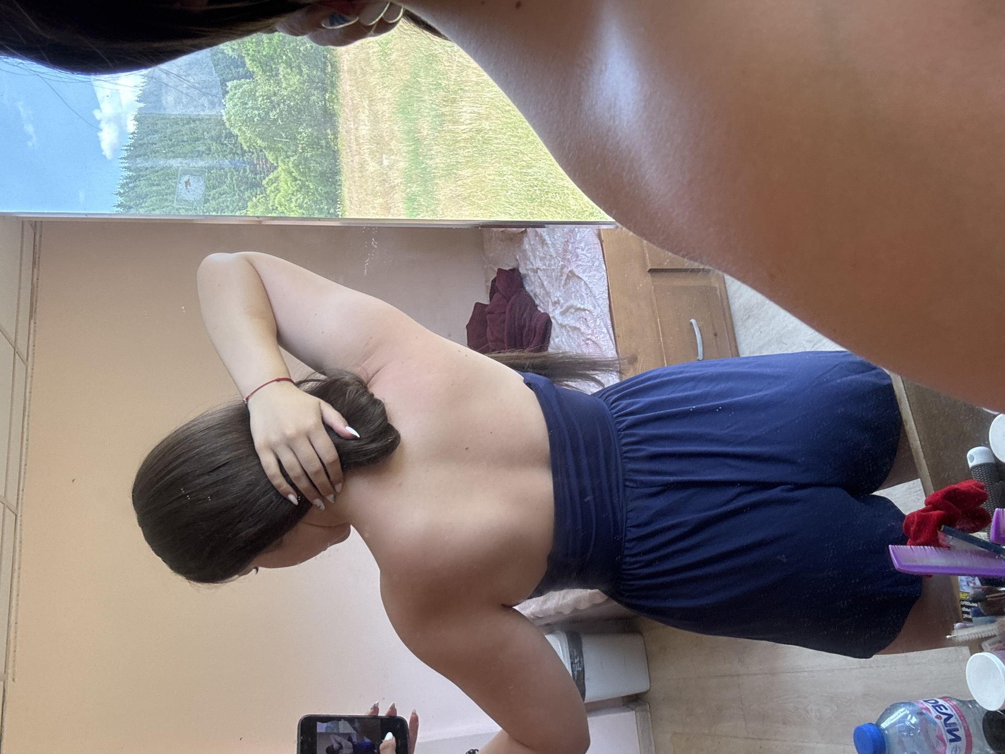 The image shows a young woman standing indoors, taking a mirror selfie. She has long, dark brown hair, and is wearing a dark blue strapless romper. Her left arm is raised and bent, with her hand resting on the back of her head. In the background, a mirror reflects a window with a view of a grassy field and trees under a blue sky. The room appears to be a bedroom, with a bed visible in the reflection. On a surface in the foreground, a plastic water bottle and various grooming items are present. The overall scene suggests a casual, personal moment captured in a residential setting.