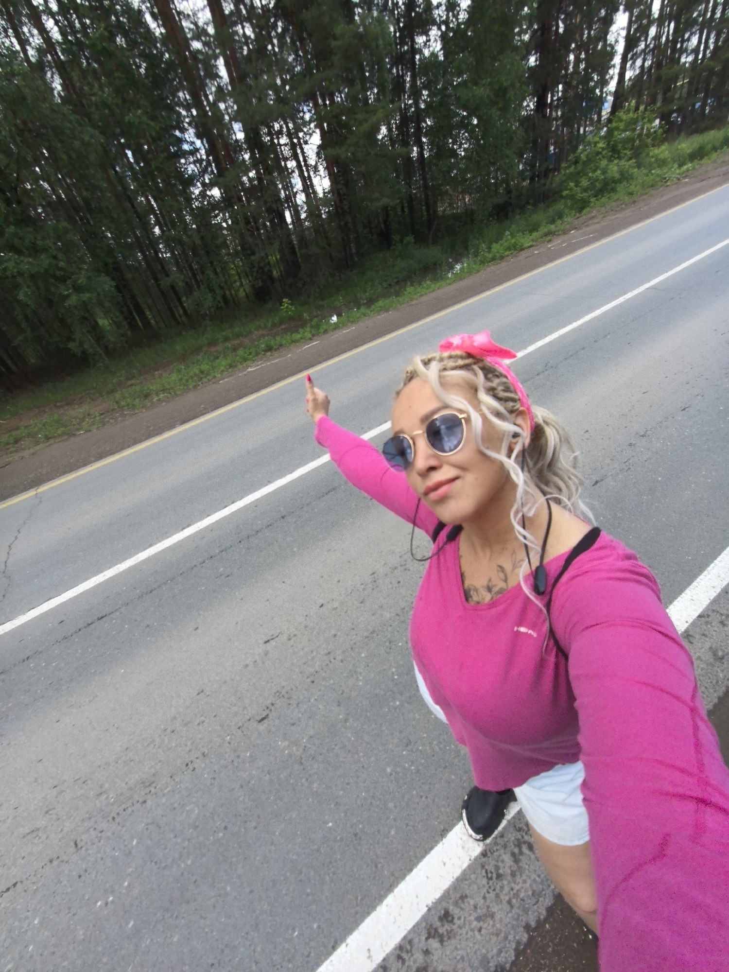 A woman with blonde braided hair and a pink headband stands on the side of a road in Noviy, Russia. She is wearing sunglasses, a bright pink long-sleeved shirt, and white shorts. Her right arm is extended with her index finger pointing upward, and she is looking at the camera. She has tattoos on her chest and is wearing headphones. The road is paved with a white line marking its edge. To the left of the road is a grassy area and a dense forest with tall trees. The sky is overcast.