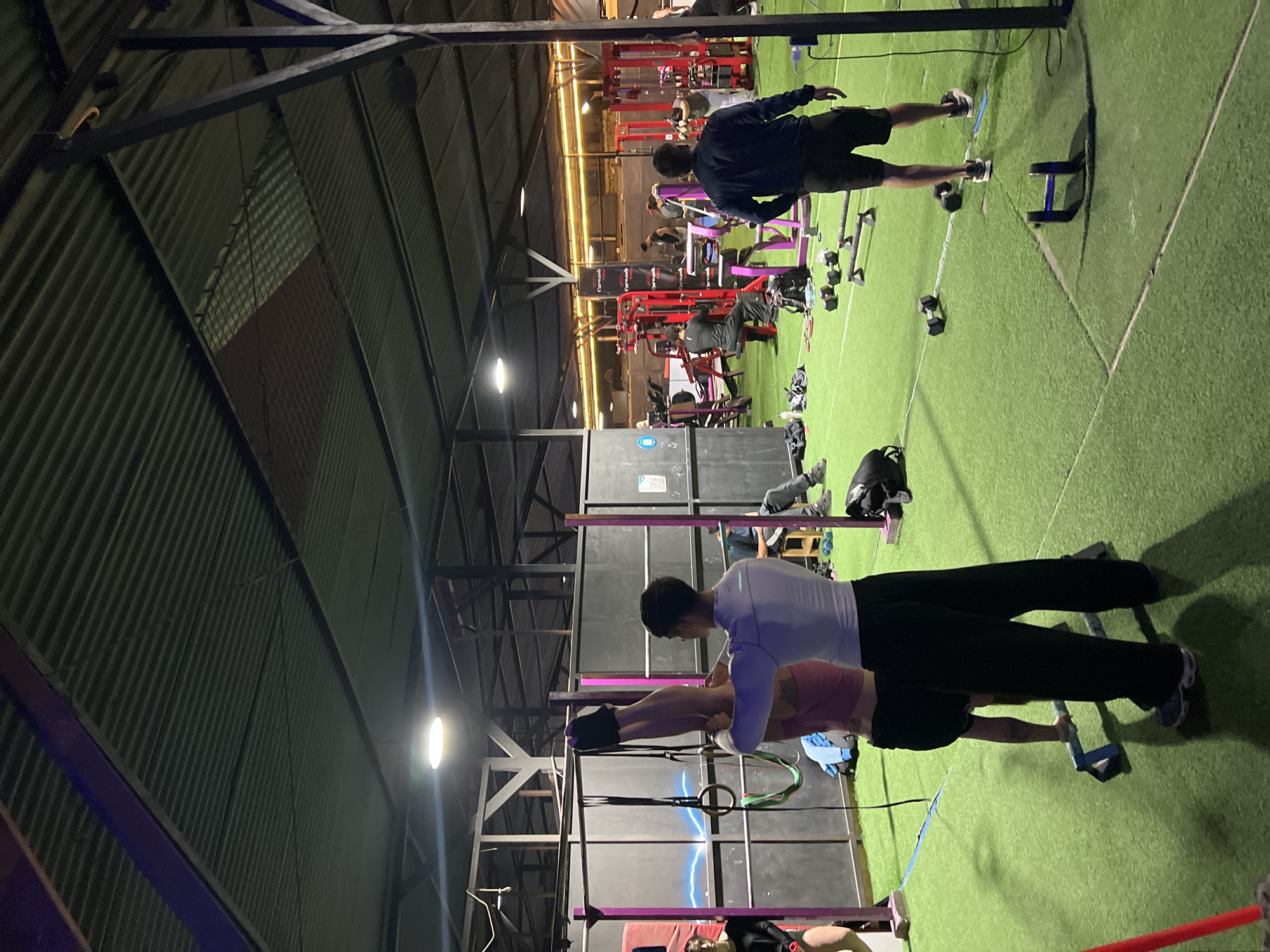 Arica, Chile: An indoor fitness facility. Individuals engage in varied exercises across artificial turf. One person helps another in a gymnastic rings drill. In the distance, machines and other people doing exercise are visible. Dumbbells are lying on the floor.