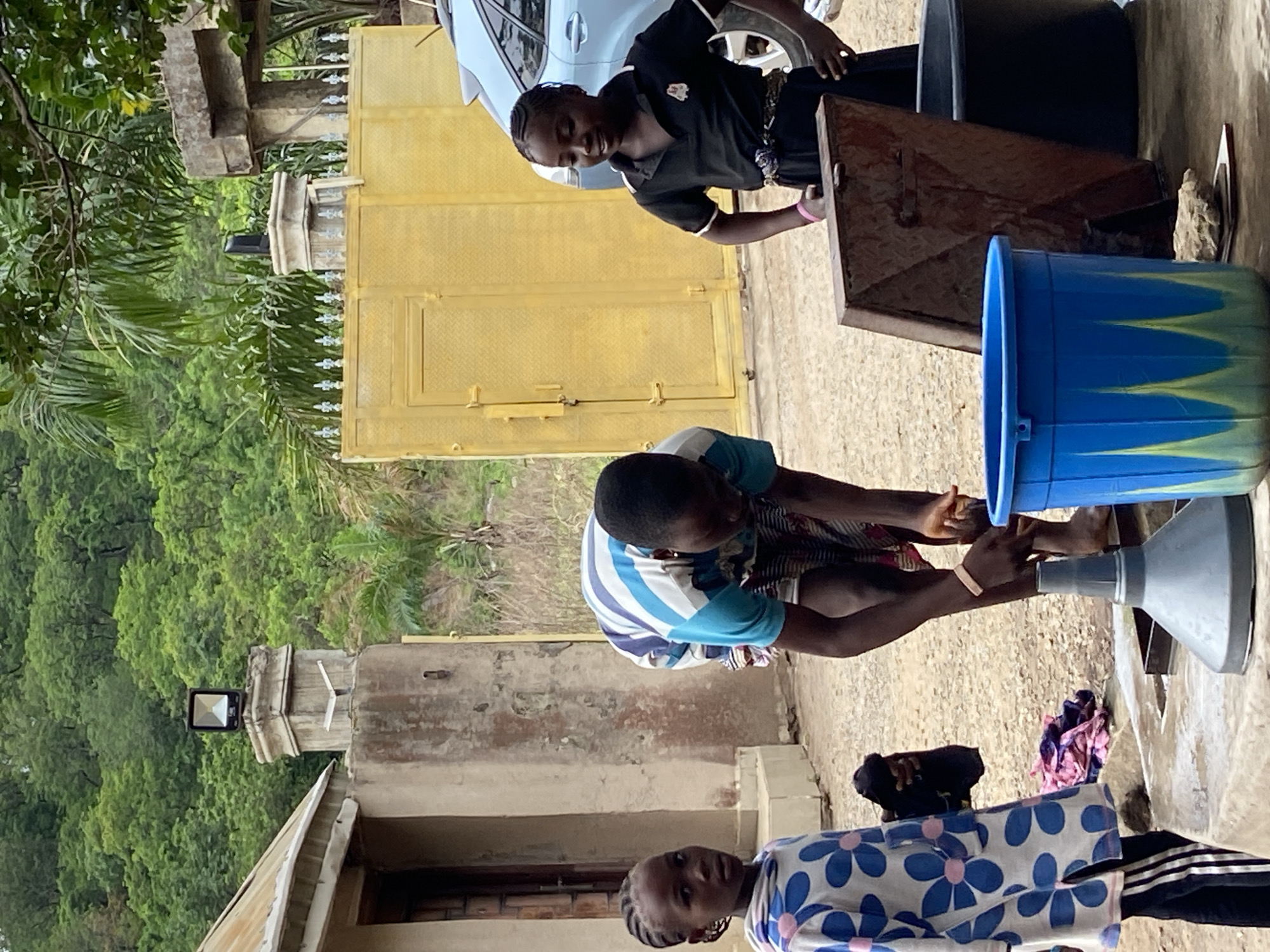 Three individuals are present in an outdoor setting. One person, wearing a black shirt, stands near a metal container and a light blue car is visible in the background. Another individual, dressed in a blue and white striped shirt, appears to be engaged in washing their hands. A blue bucket and a gray funnel are also in proximity. The third individual, wearing a shirt with blue and white floral patterns, is positioned closer to the camera. A building is visible in the lower portion of the frame, set against a backdrop of green foliage and what appears to be a metal gate. The location is Mararaba, Nigeria.