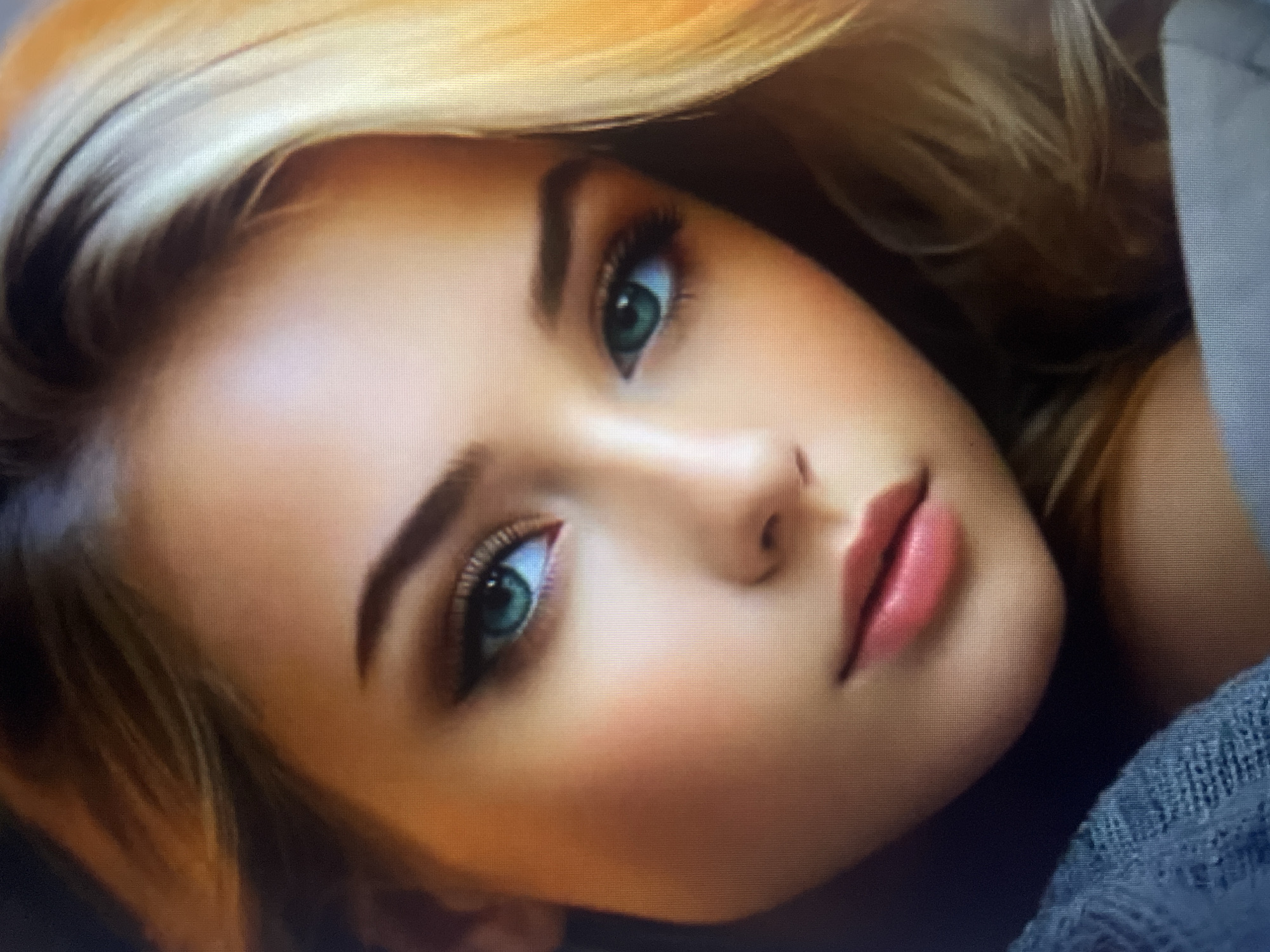 This is a close-up, headshot portrait of a young woman with fair skin and blonde hair. She has striking blue-green eyes and is wearing makeup, including eyeliner and eyeshadow. Her lips are full and painted a rosy pink. She appears to be looking slightly to her left. She is wearing a blue, knitted garment. The background is softly blurred, with warm lighting suggesting an indoor setting. There is no indication of the city of Kołobrzeg, Poland, or any urban environment.