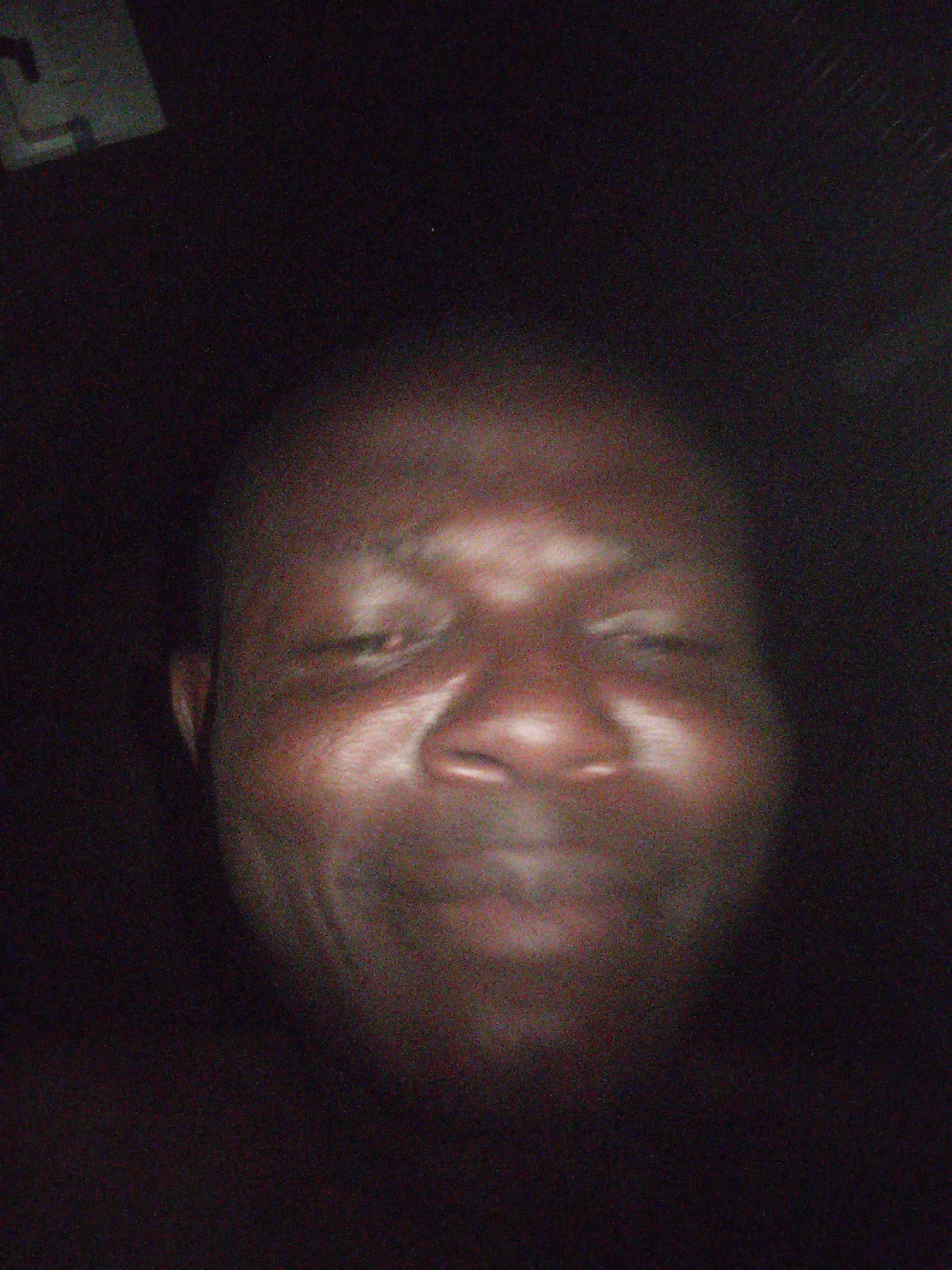 A low-resolution image shows a close-up of a Black male face, illuminated against a dark background. The subject appears to be smiling subtly. A blurred object is visible in the upper left corner.