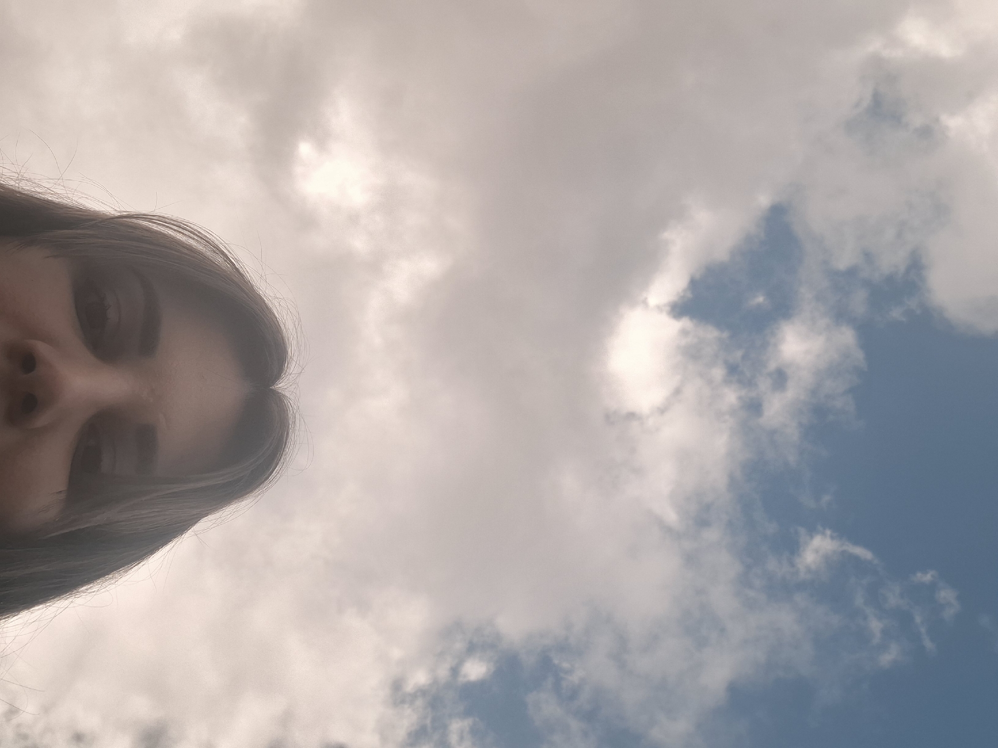 The image is taken from a low angle, looking up at a woman's face and the sky. The woman's face occupies the lower left portion of the frame. Her dark hair is visible on the left edge. The sky makes up the majority of the image, filled with white and grey clouds against a backdrop of blue. There's no information in the picture about the location being Povarenki, Russia.