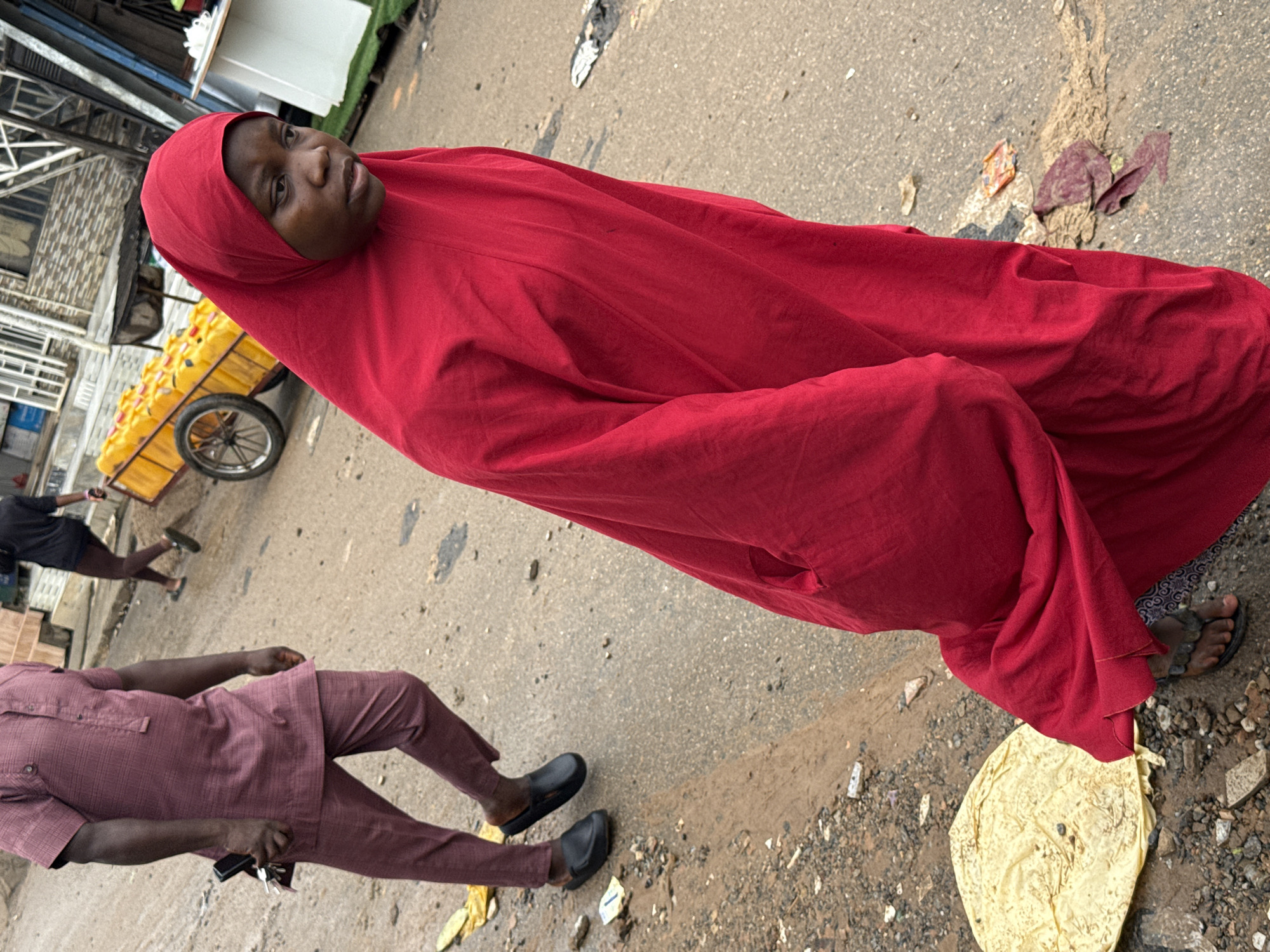 In this media file, a person is seen wearing a bright red hijab and a matching red garment. The individual is seated on the ground, with their legs tucked beneath them. To the left, another person is visible from the knees down, wearing a maroon outfit and dark slip-on shoes. This individual is holding a set of keys. In the background, a cart filled with yellow containers is being pushed by a person wearing a black outfit. The scene appears to be a street or marketplace, with a paved surface, debris, and a hint of buildings in the distance. The overall impression is one of an outdoor activity, likely during daylight hours, given the natural lighting. The subjects' expressions are not clearly discernible due to the angle and framing of the shot. The setting suggests an urban environment in Abuja, Nigeria, given the context provided.