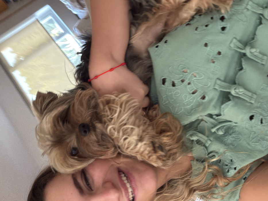 The image depicts a female individual, partially visible on the left, with light-colored wavy hair and a smiling expression, holding a Yorkshire Terrier dog. She is wearing a light green garment featuring eyelet embroidery and scalloped details across the chest area. A red string bracelet with multiple knots is present on her right wrist. The Yorkshire Terrier, identifiable by its tan and dark grey fur, small dark eyes, and pointed ears, has its head positioned centrally in the frame and looks directly at the camera. Its forepaws are visible resting on the female's chest. The background indicates an indoor domestic environment, with a window and a light-colored roller blind visible. The location is San Bernardino Tlaxcalancingo, Mexico.