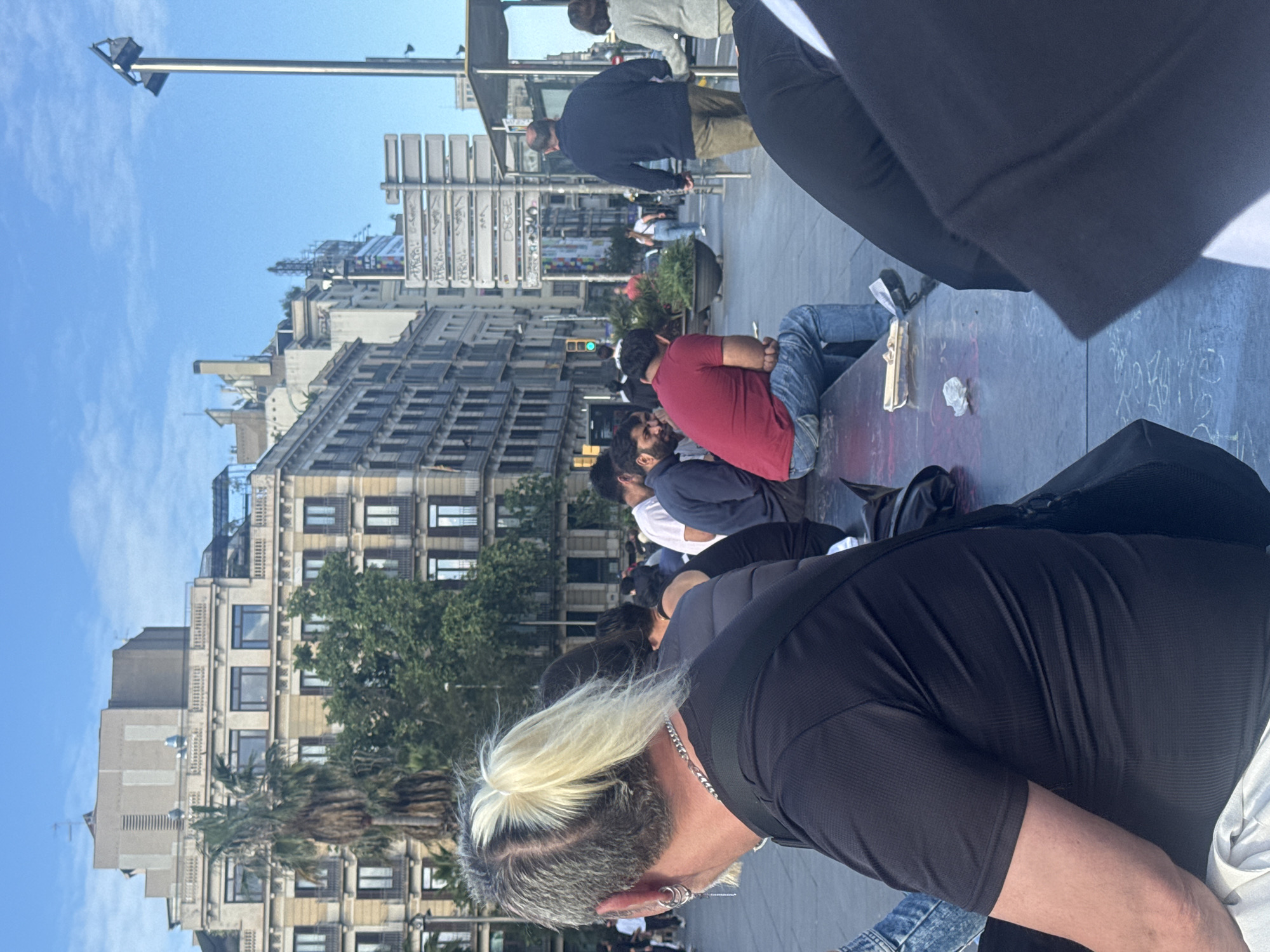 The image shows a street scene in Barcelona, Spain. Buildings line the street, and a group of people are sitting on a raised concrete platform. Some are wearing casual clothing, like jeans and t-shirts. A man in the foreground has a distinctive hairstyle, with grey hair and a blond ponytail, and is wearing a black shirt. The platform has some debris on it, and a black bag is also visible. The street appears to be a pedestrian area.