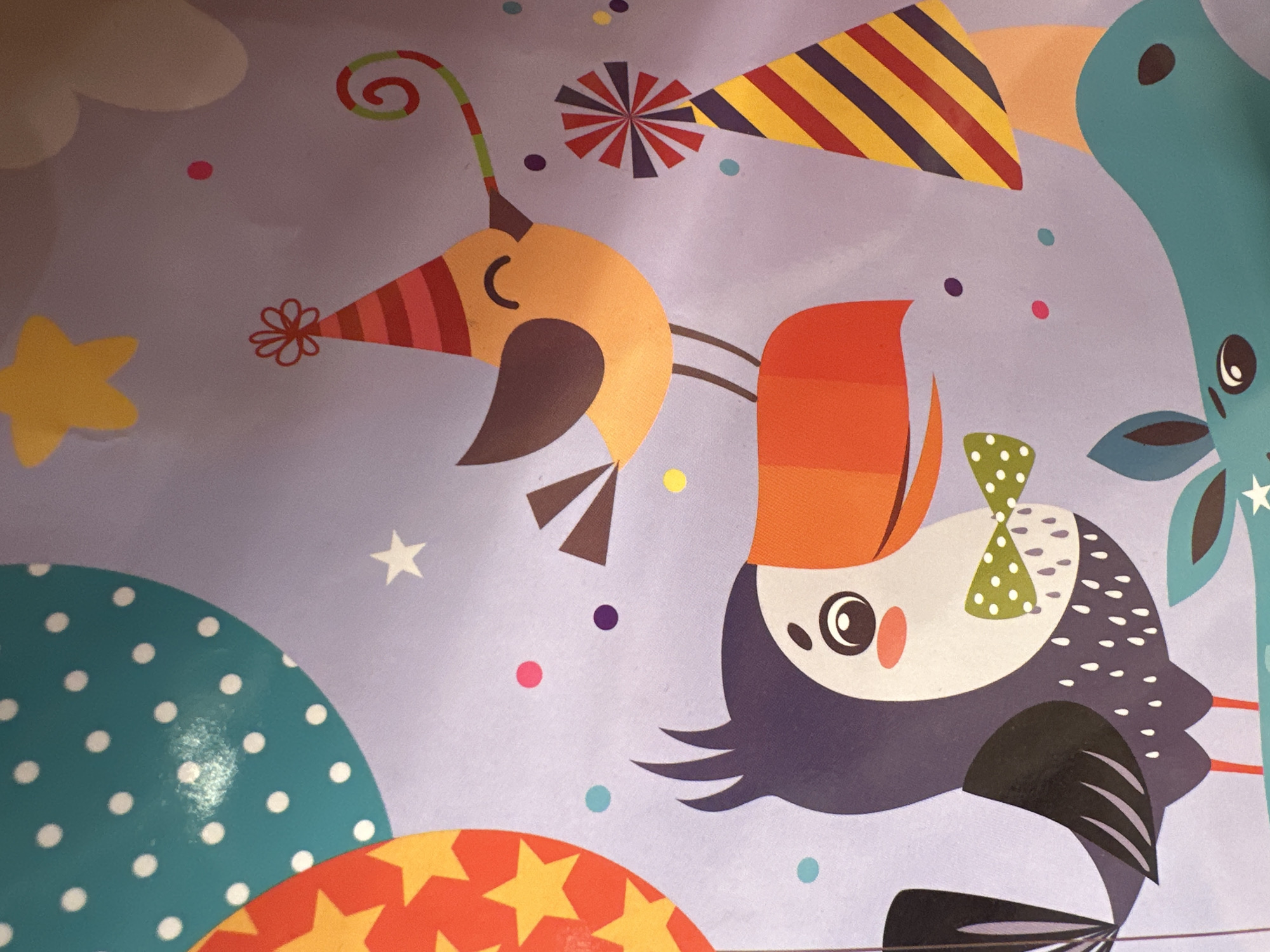 The image is a close-up, overhead shot of a playful and colorful illustration, likely on wrapping paper or a similar material. The dominant background color is a light purple.
The subjects are cartoonish birds and a giraffe. One bird, primarily orange and brown, wears a striped party hat and has a spiral-like green and red flourish extending from its hat. Another bird, predominantly purple and white with grey accents, is upside down and appears to be wearing a green bow with white polka dots. It has a large eye and a small pink cheek. A blue giraffe is partially visible on the right side of the frame, with its head and neck curved. The giraffe has large black eyes. Scattered around the figures are various festive elements: stars (one yellow, one white), colorful confetti dots (pink, purple, turquoise, yellow), and party hats (one striped in yellow, red, and purple). In the lower left and right of the frame, large, curved shapes are visible. The one on the left is teal with white polka dots, and the one on the right is red with yellow stars.
The overall scene suggests a celebration or party, possibly a birthday, given the party hats and festive decorations. The style is whimsical and cheerful. There are no discernible location cues, time of day indicators, or weather. The emotions conveyed are joy and fun. No text is visible in the image.