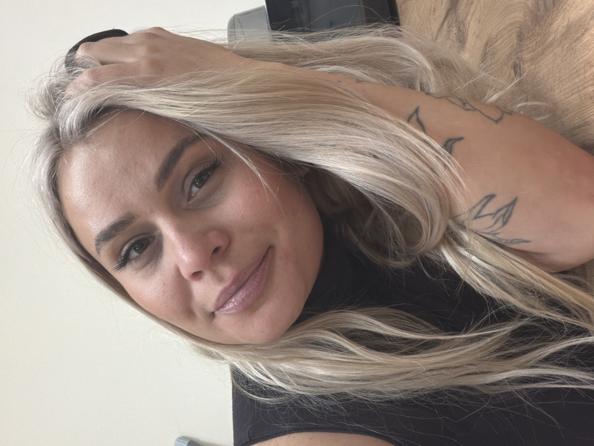 A close-up selfie shot features a woman with long, blonde hair with gray roots, and visible dark tattoos on her right forearm. Her hand is positioned in her hair. She is wearing a black top. The background appears to be a wooden surface. The city of Sarapul, Russia is not discernible from the image.