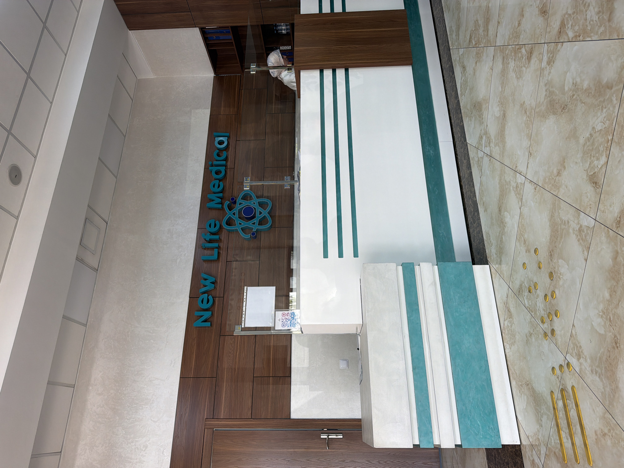 The image shows the reception area of a medical facility identified by the "New Life Medical" signage. The reception desk is modern, with a white and teal color scheme. Behind the desk, a person wearing a head covering and mask is visible, suggesting it is a healthcare setting. The walls are decorated with wooden panels and textured wallpaper. The floor is tiled with a marble-like pattern. In the foreground, yellow tactile paving and some scattered coins are visible on the floor, indicating a public space. The facility is located in Tashkent, Uzbekistan.