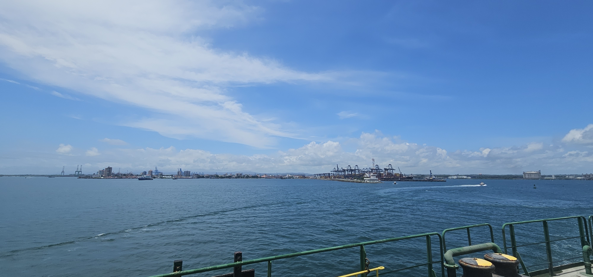 The image captures a wide view of the Panama Canal entrance area in Barrio Norte, Panama, under a partly cloudy blue sky. The foreground shows the railing of a vessel, painted green with some yellow accents. The midground is dominated by the expanse of the sea, with gentle waves and white wakes from moving boats. The coastline is visible in the distance, featuring a bustling port complex with numerous large shipping cranes, industrial buildings, and the silhouette of a city skyline. Several ships are present in the water, including container ships and smaller vessels. A small white boat is seen moving swiftly across the water, leaving a significant wake. The overall scene depicts a vibrant and active maritime hub.