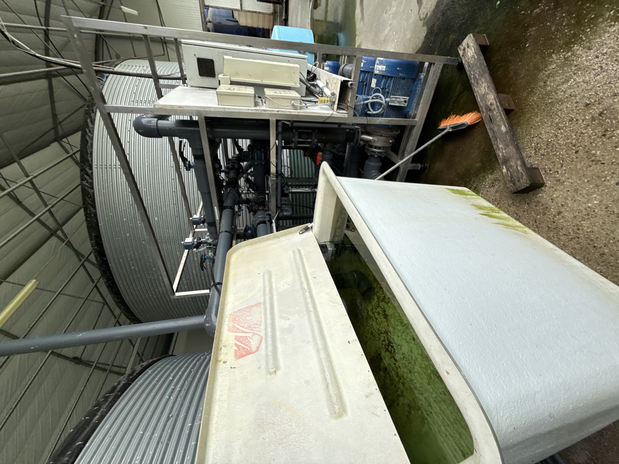 The image shows an indoor industrial aquaculture facility, likely located in Stara Zagora, Bulgaria. Several large, corrugated metal tanks are visible, along with a white, rectangular tank with a partially open lid revealing green-tinted water inside. A metal frame supports machinery, including a blue motor, a series of pipes, and electrical boxes. A broom leans against a wooden beam next to the machinery. The floor is concrete.