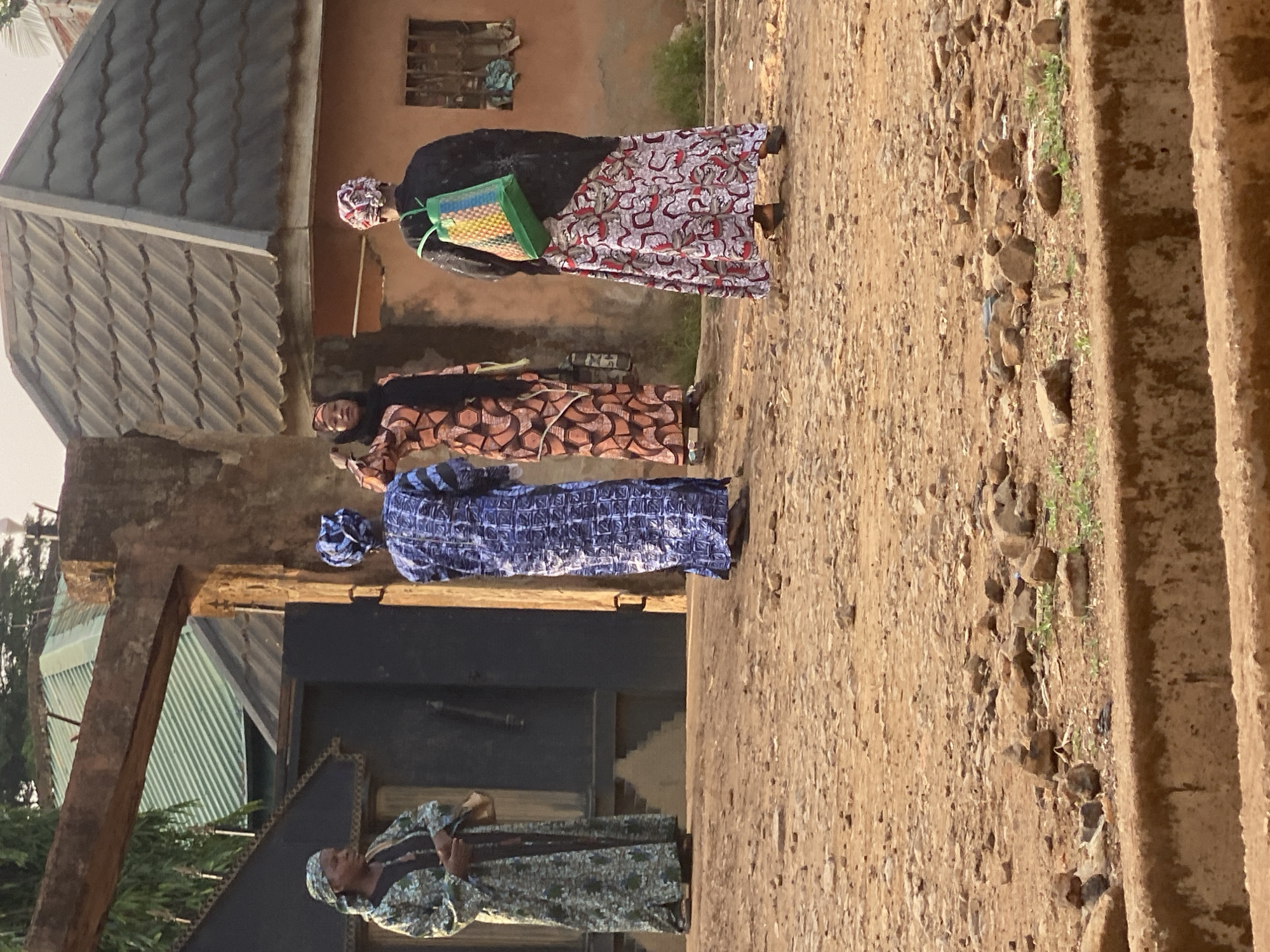 An outdoor scene in Maigizo, Nigeria, depicts four individuals, likely women, standing on a dirt and gravel path. On the far left, an individual in a light green or blue patterned long dress and head covering stands near a dark doorway. Slightly to their right, another person wears a dark blue patterned long garment and head covering. In the middle-right, a woman with a black top, an orange and brown geometric-patterned long skirt, and a headscarf faces the camera, holding a small item. On the far right, a fourth person, dressed in a black top, a white and red patterned long skirt, and a patterned head covering, carries a vibrant green woven bag on their back or shoulder.
The background includes simple residential structures: a building with a dark corrugated metal roof on the left, and a reddish-brown rendered wall with a small window further back. The path is unpaved, consisting of dirt, small rocks, and sparse greenery, leading to a rough, unpaved raised edge or steps on the right. The warm, soft lighting indicates either late afternoon or early morning. The scene captures an ordinary moment of people standing together in a rural or semi-urban environment. No text is visible in the image.