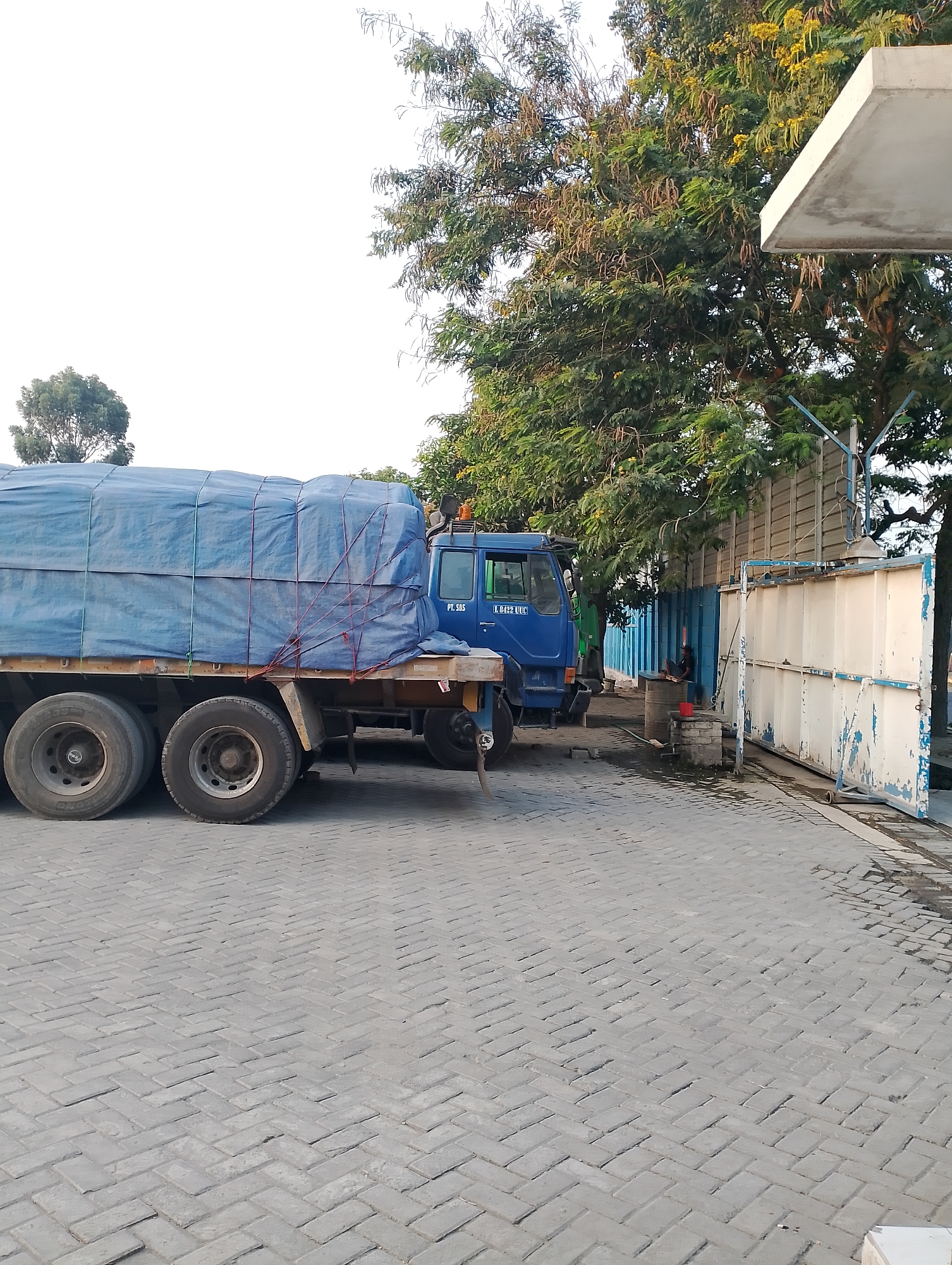A blue truck with a tarp-covered load is parked on a herringbone brick pavement in Jambean, Indonesia. The truck is positioned to the left of the frame, its rear wheels closest to the camera. A large tree with green foliage and seed pods overhangs the truck and a beige building with a concrete roof. A chain-link fence with blue and white panels separates the pavement from the building. A person is seated on a barrel near the fence, to the right of the truck. The sky is overcast and white.