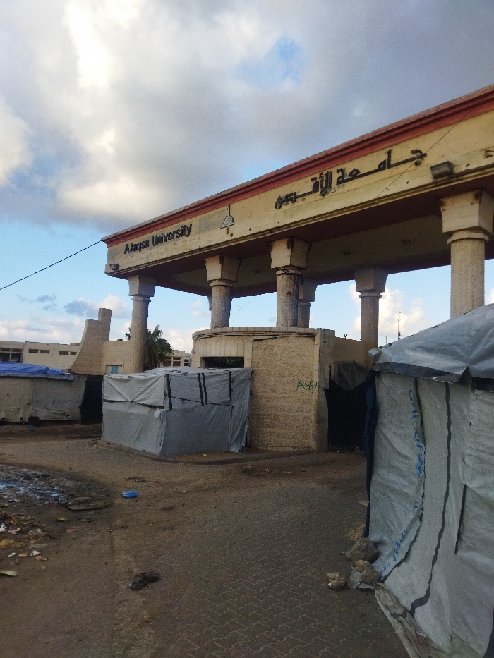 The image captures the entrance to Al-Aqsa University in Khan Yunis, Palestinian Territory, under a partly cloudy sky. The main subject is a large, ornate gateway with multiple pillars supporting a broad architrave. The university's name is prominently displayed in English as "Alaqsa University" and in Arabic as "جامعة الأقصى". A small, triangular emblem or logo is also visible on the facade near the English text.
Directly beneath the main archway, a smaller, block-built structure serves as a potential gatehouse or checkpoint, with visible graffiti on its side, possibly reading "A69" or "AG9". The area in front of and immediately surrounding the university entrance appears to be a makeshift encampment. Several temporary shelters or tents, constructed from grey and white tarpaulin sheets, are visible on both sides of the gateway. One tent in the foreground on the right side shows a faint "UNICEF" logo.
The ground consists of a mixture of dirt, mud, and broken pavement, with scattered debris and what appears to be litter. In the background, to the left of the main entrance, parts of other buildings, presumably belonging to the university, can be seen. The overall scene suggests a blend of established infrastructure with temporary, possibly displaced, living arrangements, indicative of difficult conditions in the region. The lighting indicates it is daytime, with soft light filtering through the clouds. No people are visible in the frame.