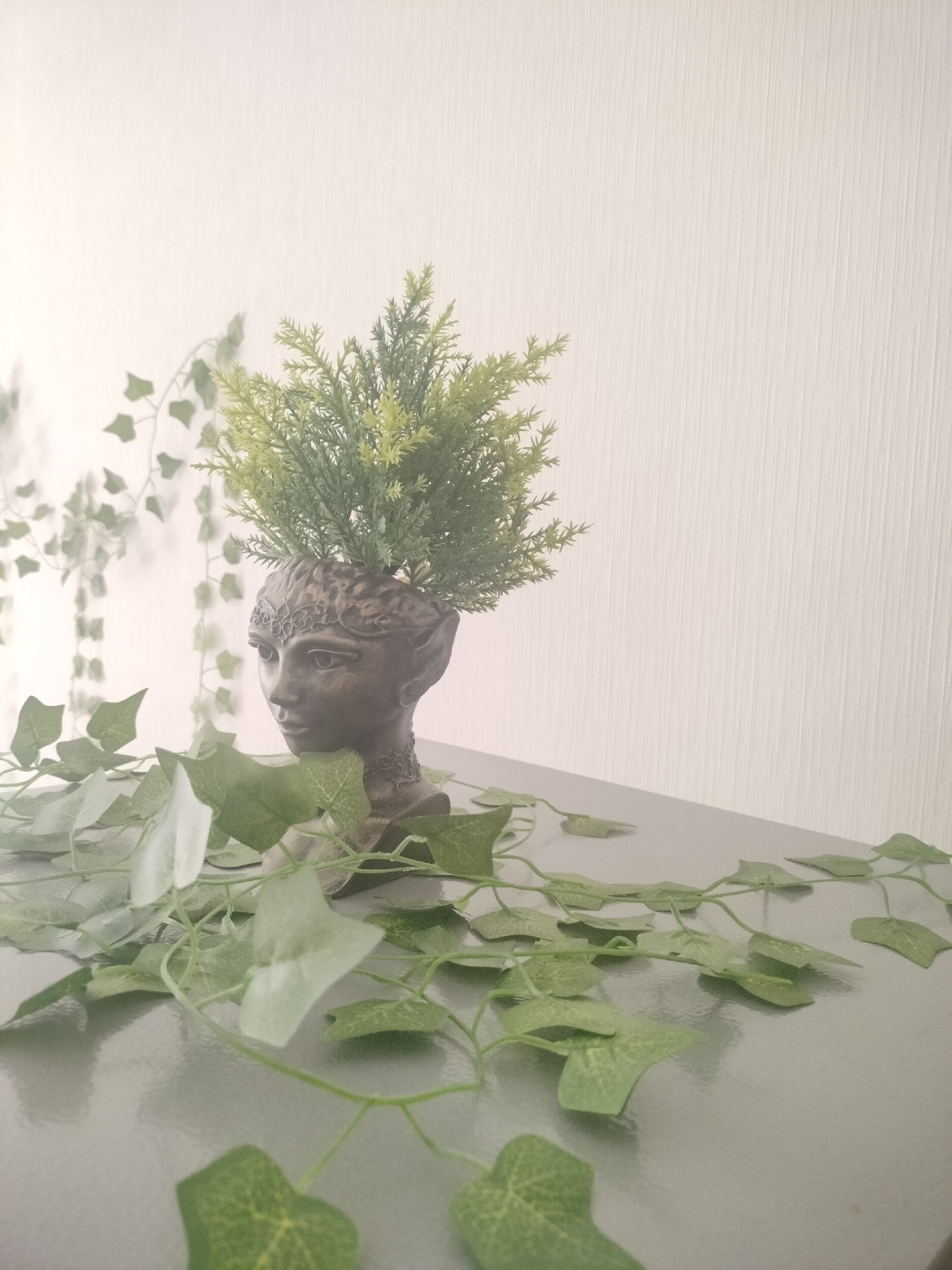 The image shows a bust of a woman with pointed ears, likely a decorative planter, filled with a green, bushy plant. The bust is positioned on a grey, reflective surface, possibly a table or shelf, in front of a textured, off-white wall. Green artificial ivy vines are draped around the bust and across the surface. The lighting is soft and diffused, casting subtle shadows. There is no visible indication of the location in Lyubertsy, Russia, or any people or actions.