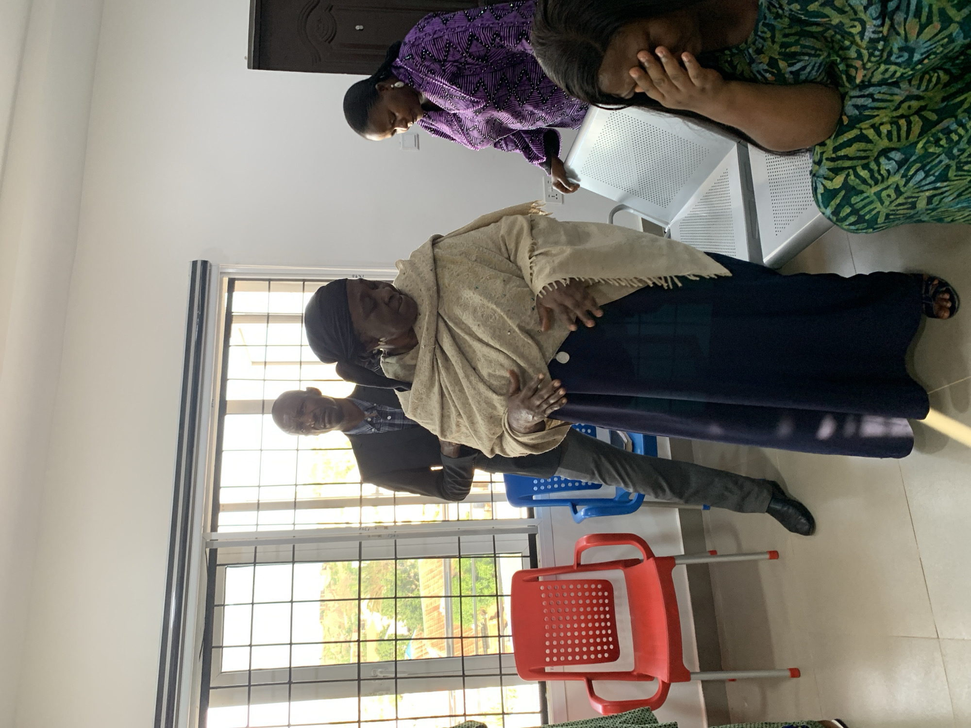 This media file shows a group of people in what appears to be a waiting area or reception. The image is oriented vertically, so the people are standing and sitting sideways relative to the viewer.
In the foreground, an elderly woman is seated and draped in a light-colored shawl. She is looking down. To her left, a man in a dark suit and collared shirt is standing, looking towards the window.
Further back, a woman in a purple, intricately patterned garment is leaning forward, her gaze also directed downwards. To her right, another woman, wearing a green and yellow patterned outfit, has her hand covering her face, suggesting distress or grief. She is seated.
The scene appears to be indoors, with large windows suggesting a daytime setting. The background shows a light-colored wall and chairs, including a red plastic chair in the foreground and some white metal chairs. The overall atmosphere seems somber, with the woman covering her face hinting at a potentially sad event, such as a funeral or a difficult situation. There is no visible text in the image.
