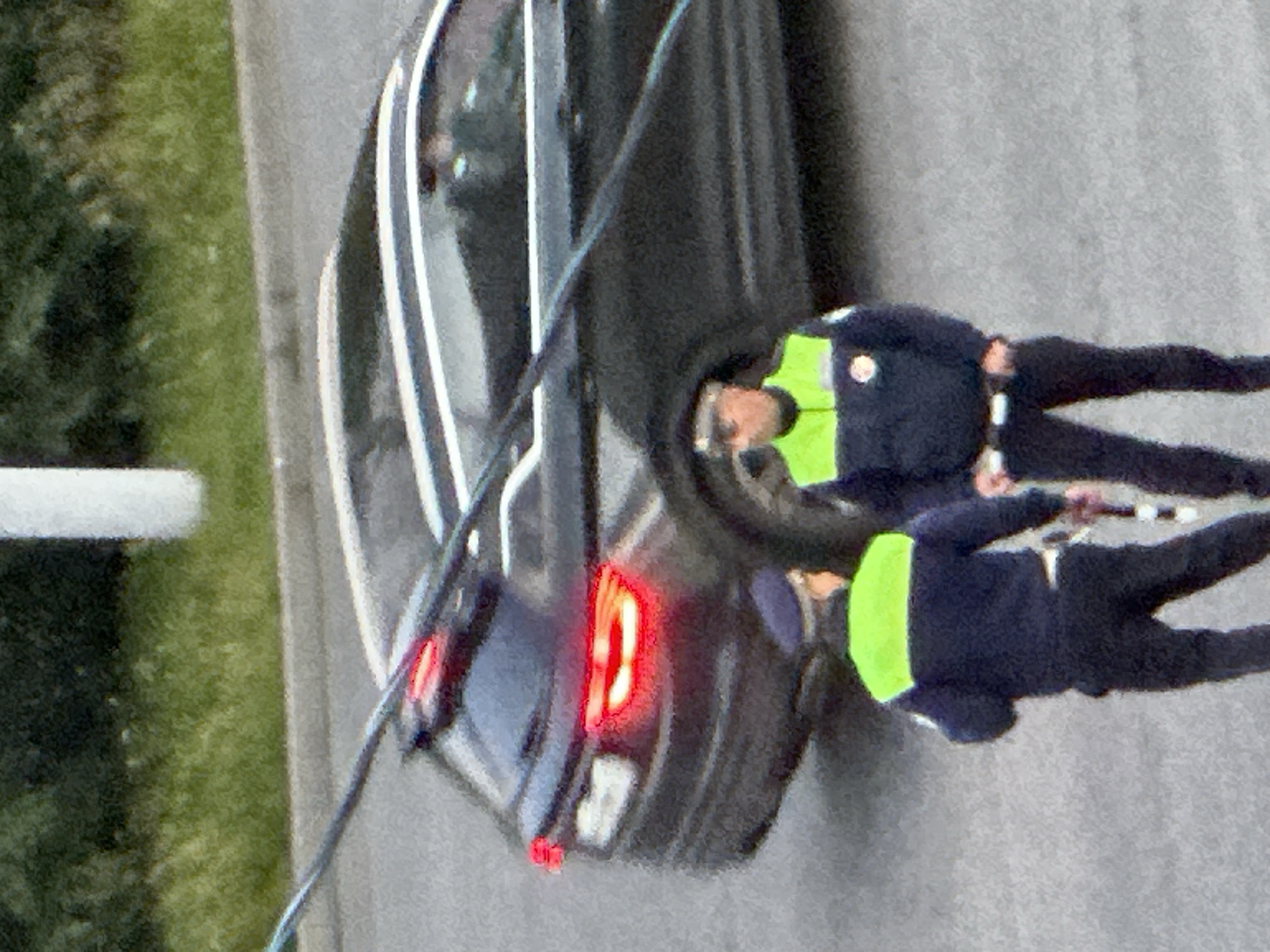 Two police officers in dark blue uniforms with high-visibility green vests are standing on a paved surface, possibly a road or parking lot. The officer on the left is facing away from the camera, while the officer on the right is facing mostly towards the camera. Both officers are wearing dark pants and caps. Behind them, a dark-colored SUV is parked, with its brake lights illuminated. A green, grassy area with some bushes is visible in the background, along with a white cylindrical structure. A dark, diagonal wire or cable traverses the upper portion of the image. The scene appears to be outdoors, under daylight conditions. The image is grainy and has low resolution, making it difficult to discern specific facial features or insignia. The location is identified as Podolsk, Russia.