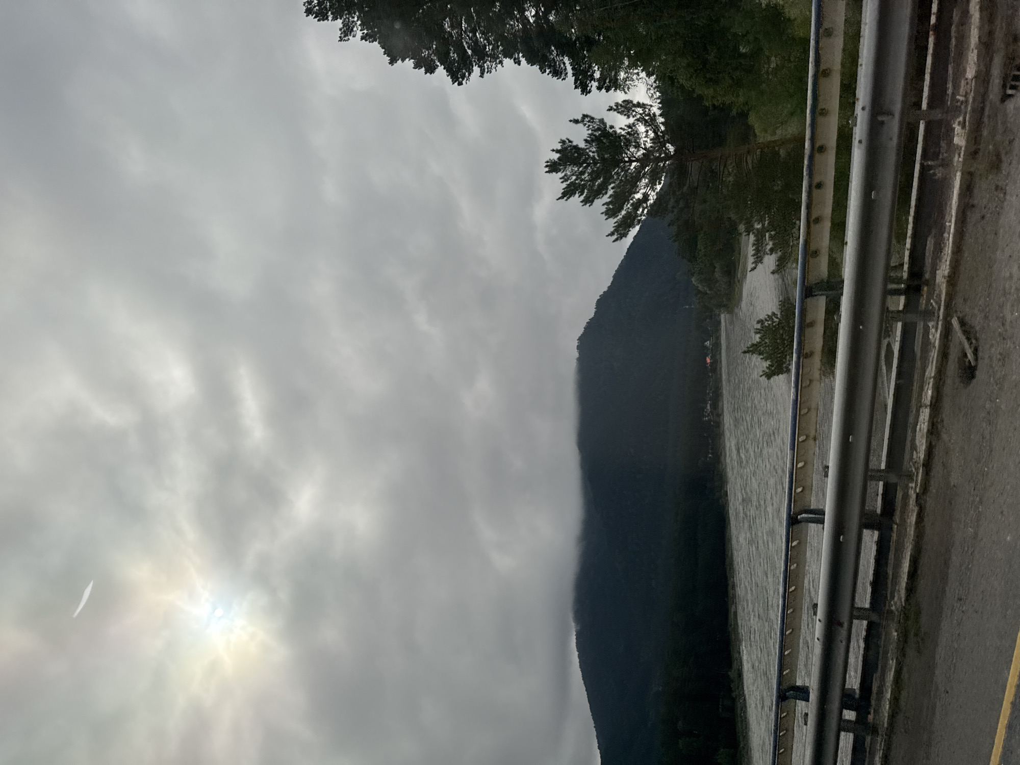 The image depicts a scenic view of a river flowing through a mountainous landscape, possibly in or near Ust'-Sema, Russia. The sky is overcast with prominent cloud formations. The sun is partially visible through the clouds, casting a soft light. A thin, white contrail is visible in the upper left portion of the sky.
In the foreground, there is a road with a guardrail. The guardrail is made of metal and appears to be supported by posts. The road surface is asphalt, with a yellow line marking one edge.
Beyond the road and guardrail, a wide, grey river flows from left to right across the image. The water appears somewhat turbulent. The riverbanks are lined with trees, predominantly evergreen.
In the middle ground, a dark, forested mountain rises from the far side of the river. The peak of the mountain is partially obscured by low-hanging clouds. Small structures, possibly houses, can be seen nestled among the trees on the riverbank in the distance to the left.
In the right mid-ground and background, more evergreen trees are visible, some of them quite tall, partially obscuring the sky and mountain. The overall atmosphere is subdued due to the cloudy weather, suggesting a cool or possibly damp environment.