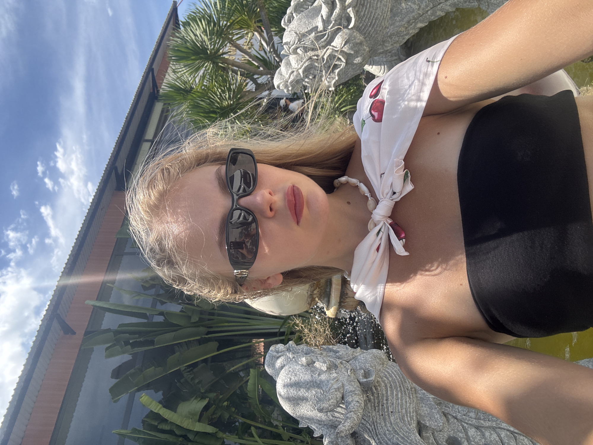 A young woman with blonde hair is taking a selfie outdoors. She is wearing a black bikini top and a white bandana with a cherry pattern tied around her neck. She is also wearing black sunglasses. The background shows a building with a corrugated roof, palm trees, and a stone statue that appears to be a dragon or mythical creature. There is a body of water in the lower right corner of the image. The sun is shining brightly, casting shadows and creating lens flare. The time of day appears to be midday, based on the strong sunlight. The woman's lips are painted red, and she is looking at the camera with a slight smile. The overall mood is casual and summery, suggesting a vacation or a day spent by the water. The sunglasses have "GUCCI" written on them. The setting is likely a resort or a private residence in Tuy An Nam, Vietnam, given the tropical foliage and architectural style.