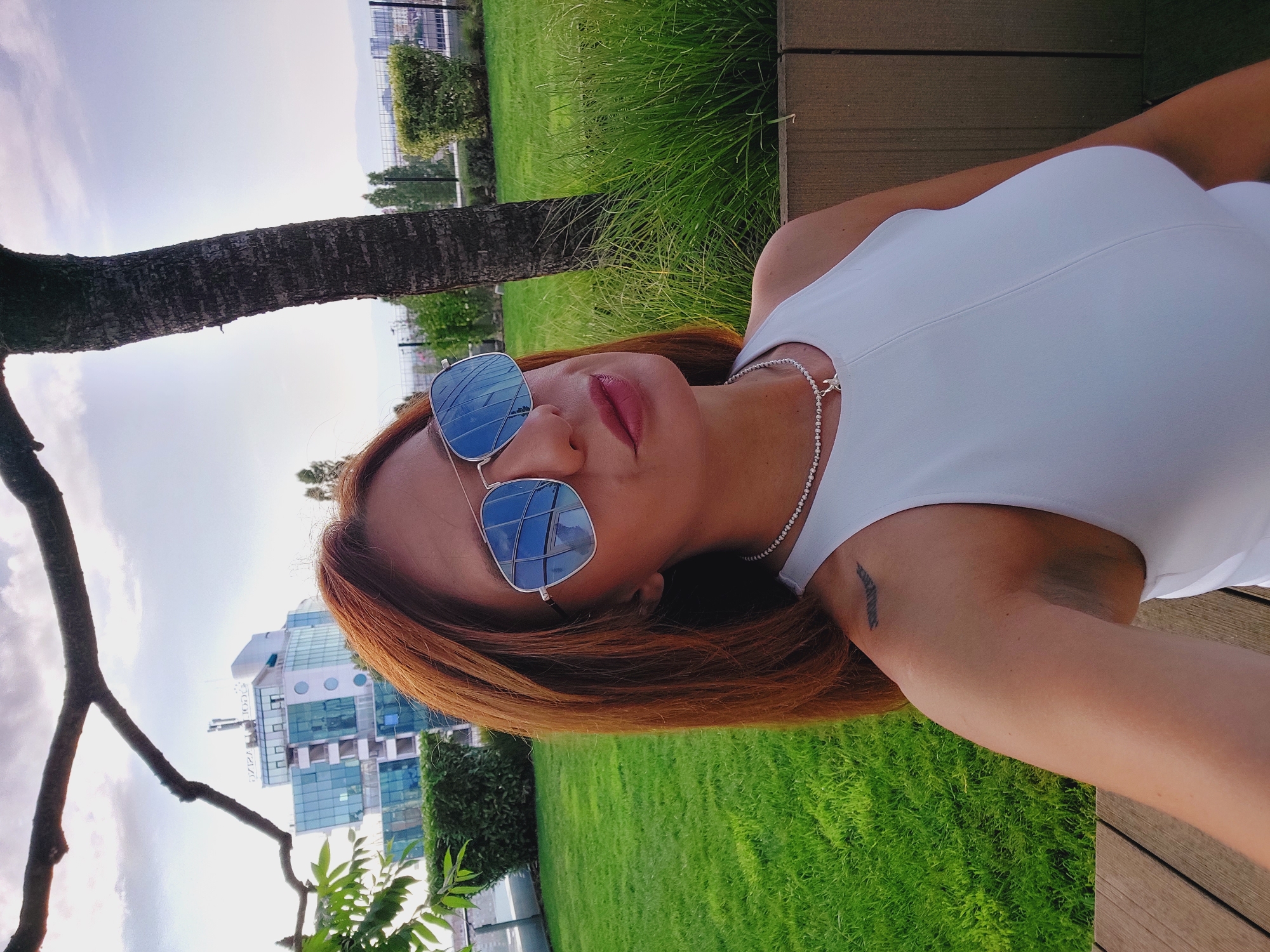 In an outdoor setting in the city of Sofia, Bulgaria, a woman with auburn hair and sunglasses is captured in a selfie. She is wearing a white sleeveless top and a delicate necklace with a pendant. Her left shoulder bears a tattoo of a wing. Behind her, a lush green lawn is visible, bordering a modern building with a glass facade. A tree trunk stands to the right of the woman, and a branch from another tree frames the upper portion of the image. The sky above is partly cloudy. The scene appears to be a rooftop garden or a terrace area, with wooden decking in the foreground.