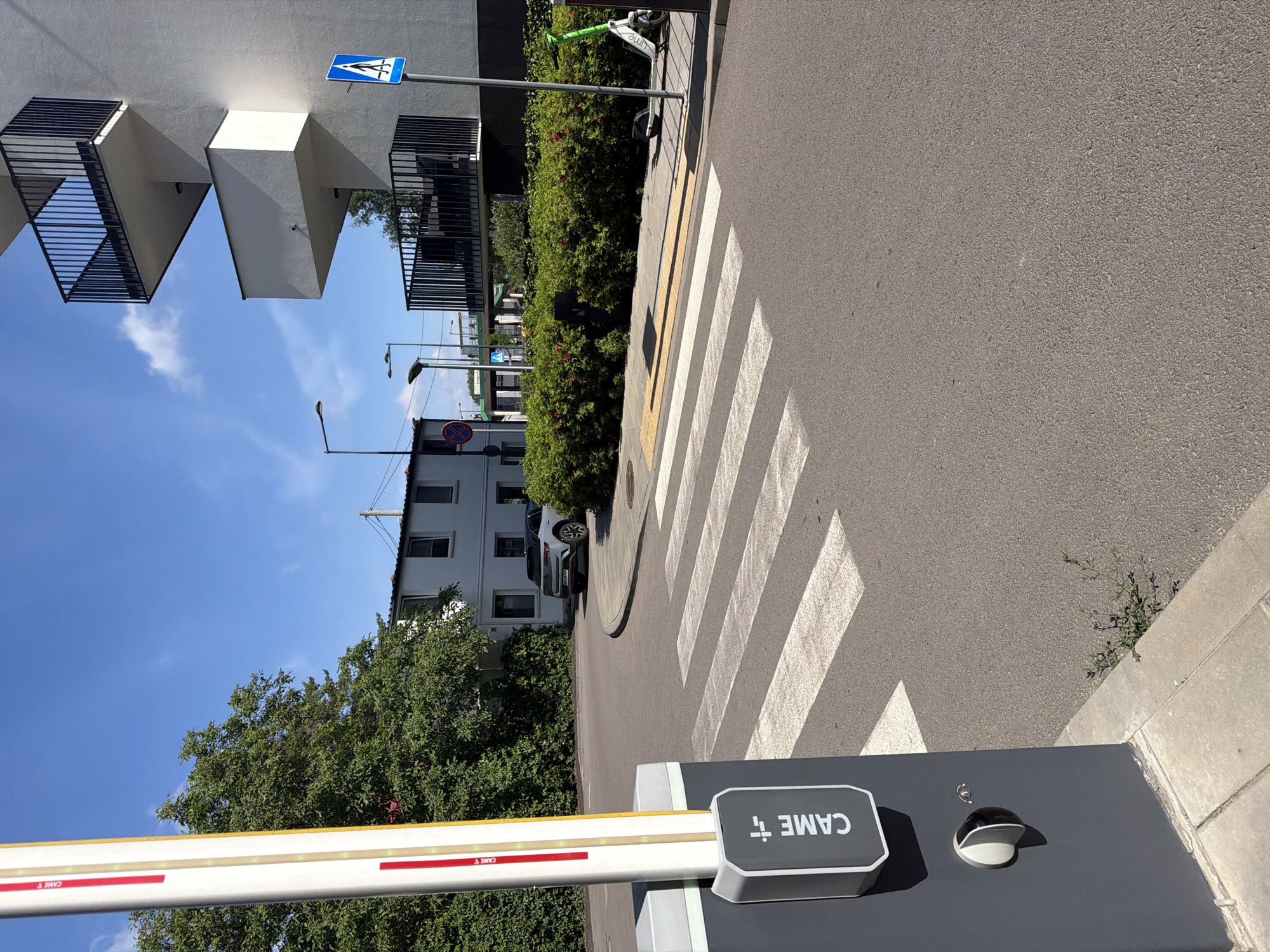 An eye-level outdoor medium shot captures a barrier gate and a crosswalk on a sunny day. The barrier gate, a tall, gray structure with a white and red striped arm, is in the foreground to the left. A gray circular button is visible on the side of the gate. Across the street, a white and yellow crosswalk stretches from right to left. Beyond the crosswalk, a white SUV is parked on the right side of the street, and a line of dense green bushes borders the sidewalk. In the background, a modern white building with dark balconies stands tall. The sky is bright blue with a few scattered clouds. A pedestrian crossing sign is visible on a pole to the right of the building. A green electric scooter is parked near the bushes on the right.