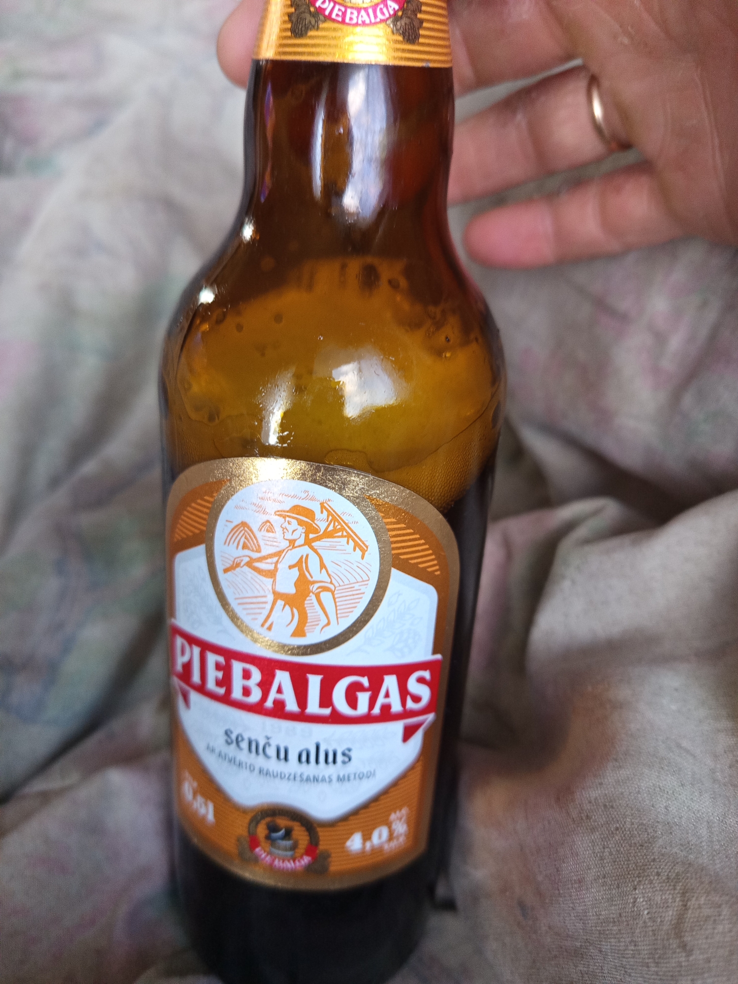 A person's hand, wearing a ring on the ring finger, is holding a brown glass bottle of "Piebalgas Senču alus." The bottle has a label with an illustration of a farmer with a scythe and haystacks, and text in Latvian indicating it's a "senču alus" (elder's beer) brewed using a traditional method. The alcohol content is listed as 4.0%. The bottle is partially filled with a dark amber liquid and appears to be sitting on a textured surface, possibly fabric. The lighting suggests an indoor environment.