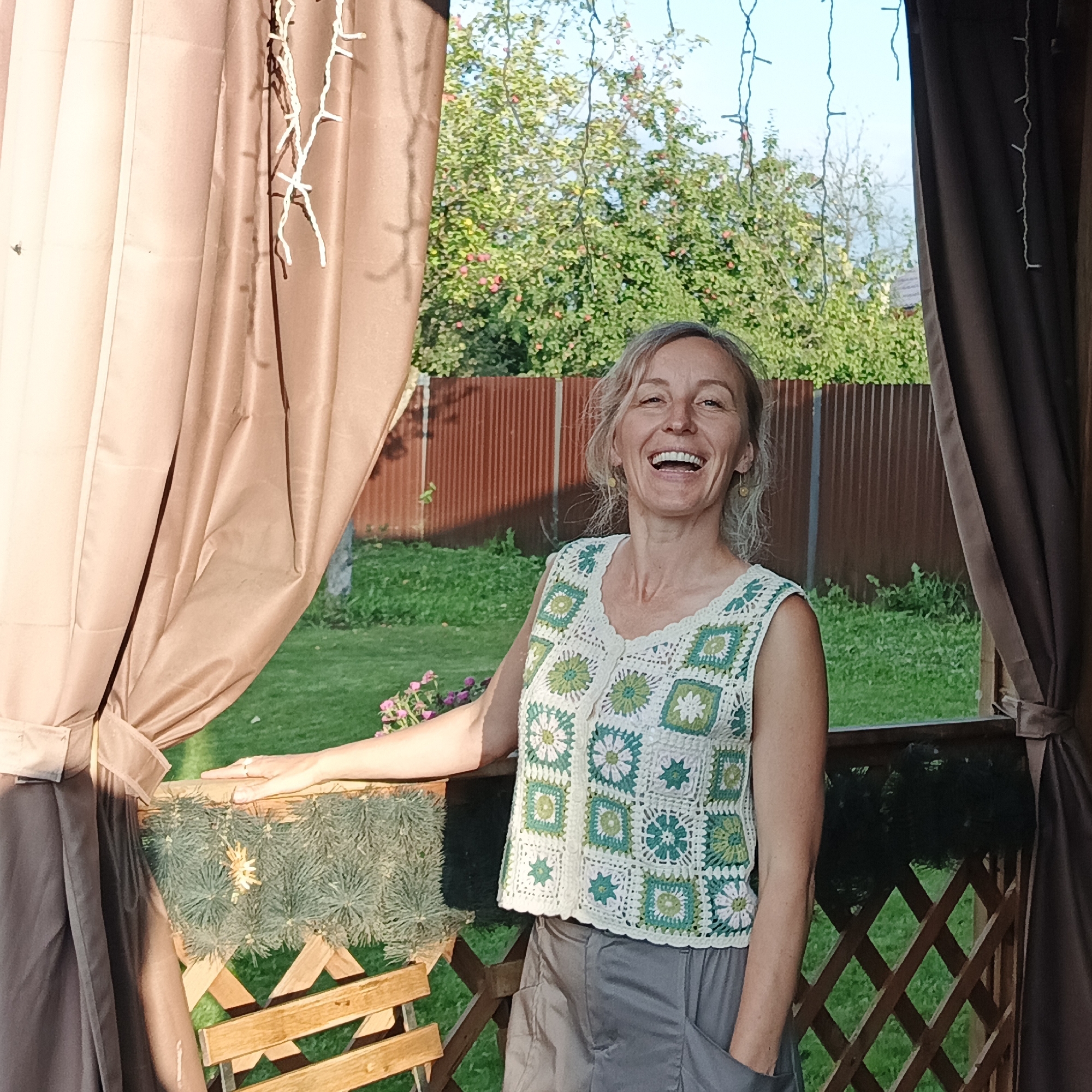 A medium shot captures a laughing woman standing on a wooden deck, framed by a brown tarp on the left and a brown curtain on the right. The woman, with blonde hair and fair skin, wears a white and green crocheted sleeveless top over grey cargo pants. She is leaning on a wooden railing decorated with green pine branches and a star ornament. Behind her, a green lawn and a row of reddish-brown corrugated metal fencing are visible, with lush green trees and a blue sky forming the background. The scene appears to be set in a backyard or a gazebo. The woman's body language and open mouth suggest genuine amusement. The lighting indicates it is daytime, likely late afternoon or early evening given the warm tones.