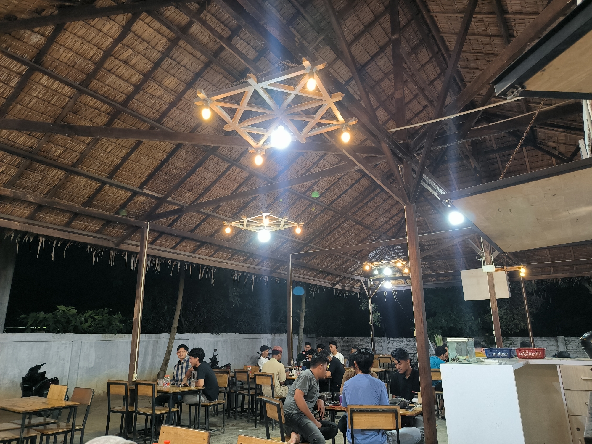 The image depicts the interior of an open-air restaurant in Cempeudak, Indonesia. The restaurant has a thatched roof supported by wooden beams, and is illuminated by decorative light fixtures. Several people are seated at tables, appearing to be engaged in conversation and dining. The restaurant is located near a wall and vegetation can be seen in the background.