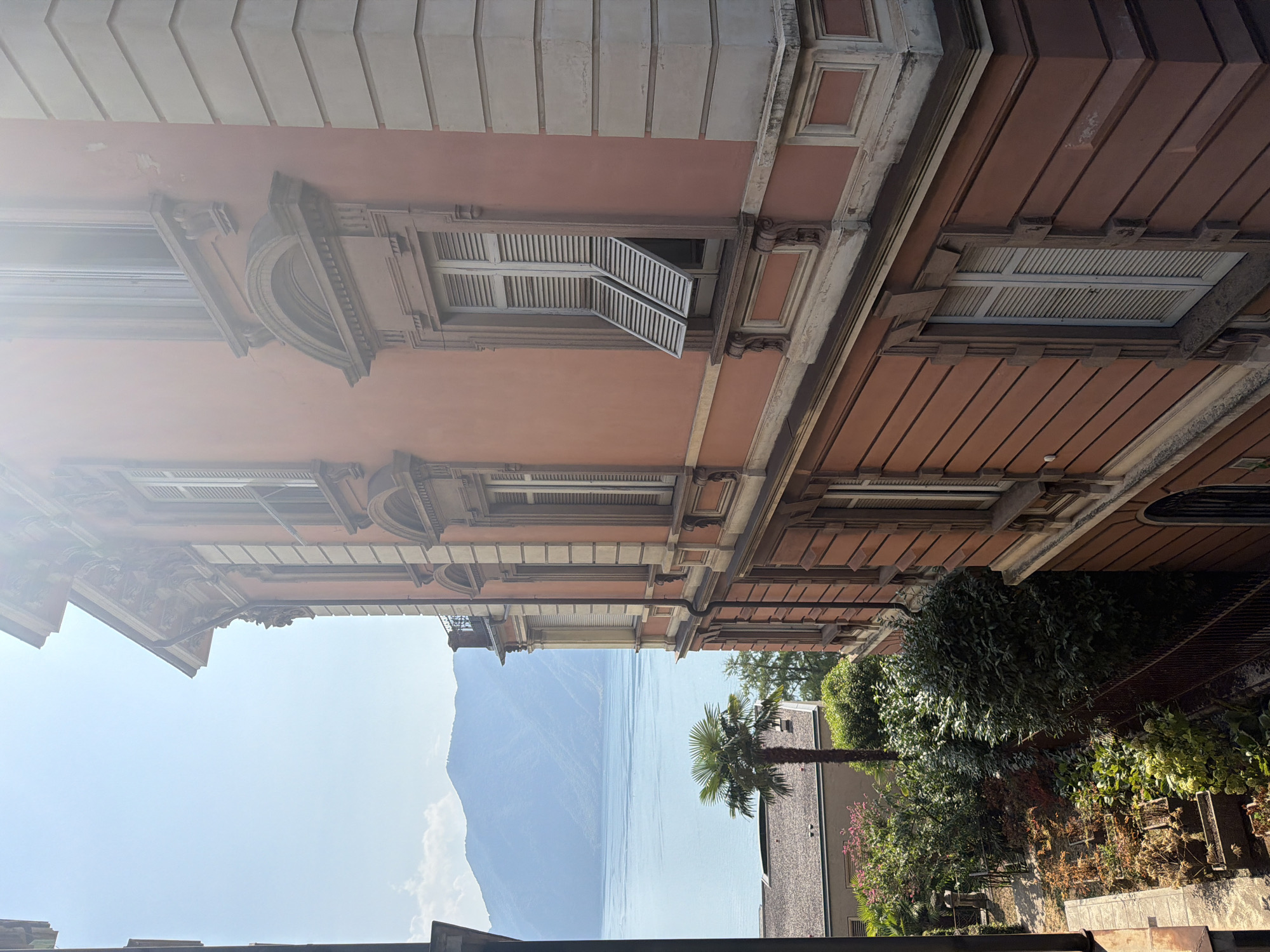 This image captures a view from a building in Lugano, Switzerland, overlooking a serene lake and mountains. The foreground is dominated by the facade of a building with warm, salmon-pink walls and ornate architectural details around its windows. Several windows are visible, some with white shutters partially open, allowing sunlight to filter through. The building's exterior features horizontal lines and decorative moldings.
To the left, a lush green garden is visible, with a variety of plants and a tall palm tree. In the distance, the expansive, calm lake stretches out, reflecting the clear blue sky. Majestic, hazy mountains rise beyond the lake, their slopes covered in vegetation. The scene is bathed in bright sunlight, creating a slightly hazy atmosphere. A sliver of another building is visible on the far left edge, indicating an urban setting. The overall impression is one of tranquil beauty, combining elegant architecture with the natural splendor of the Swiss landscape.