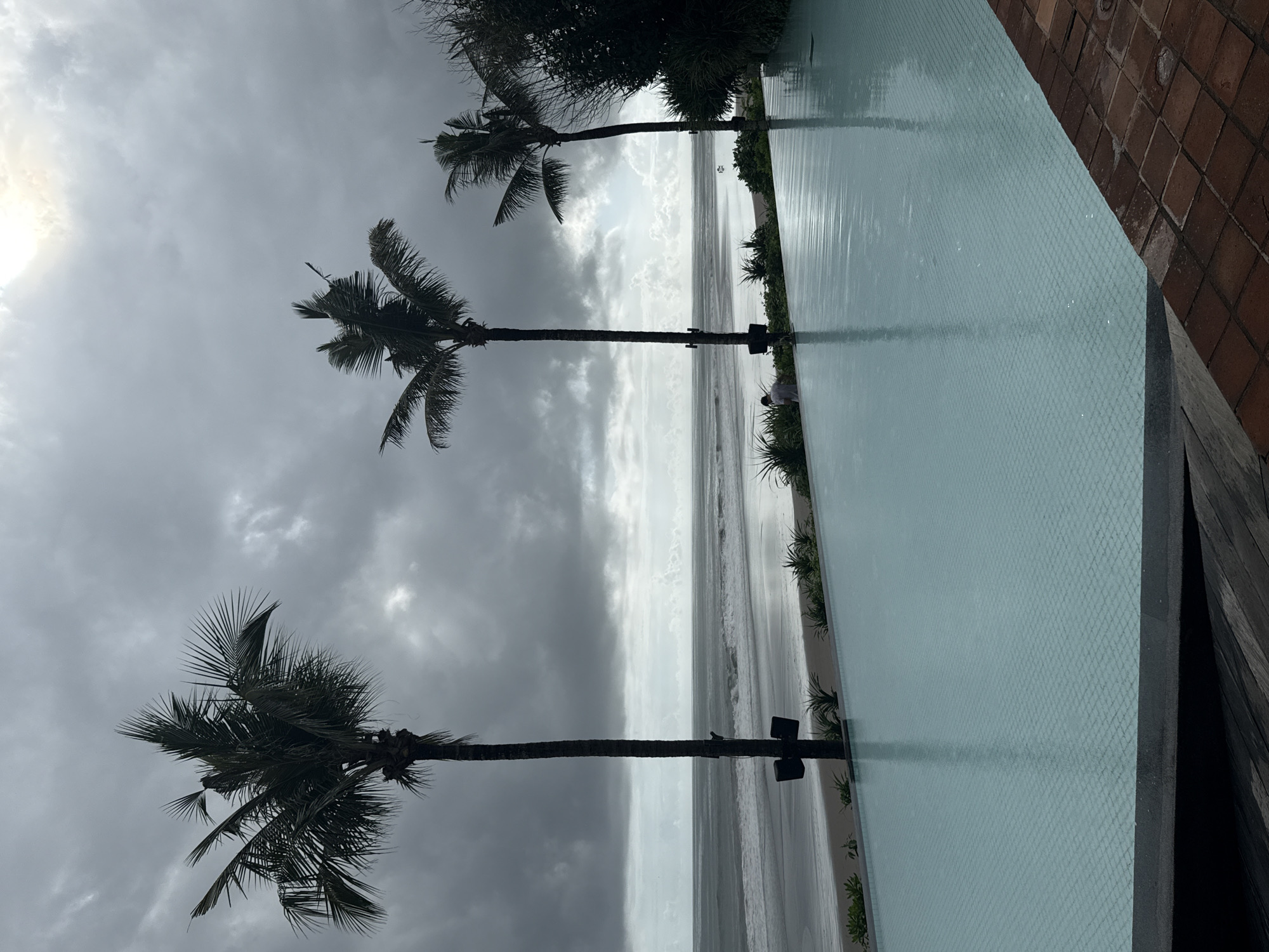 The image shows a view from a resort in North Kuta, Indonesia. Three palm trees are visible against a cloudy sky, with the sun barely peeking through. The trees appear to stand next to a pool, which reflects the overcast sky. Beyond the pool, a beach with waves can be seen. The pool's edge is adjacent to a brick-paved area, indicating the resort setting. The overall scene suggests a tropical location experiencing inclement weather.