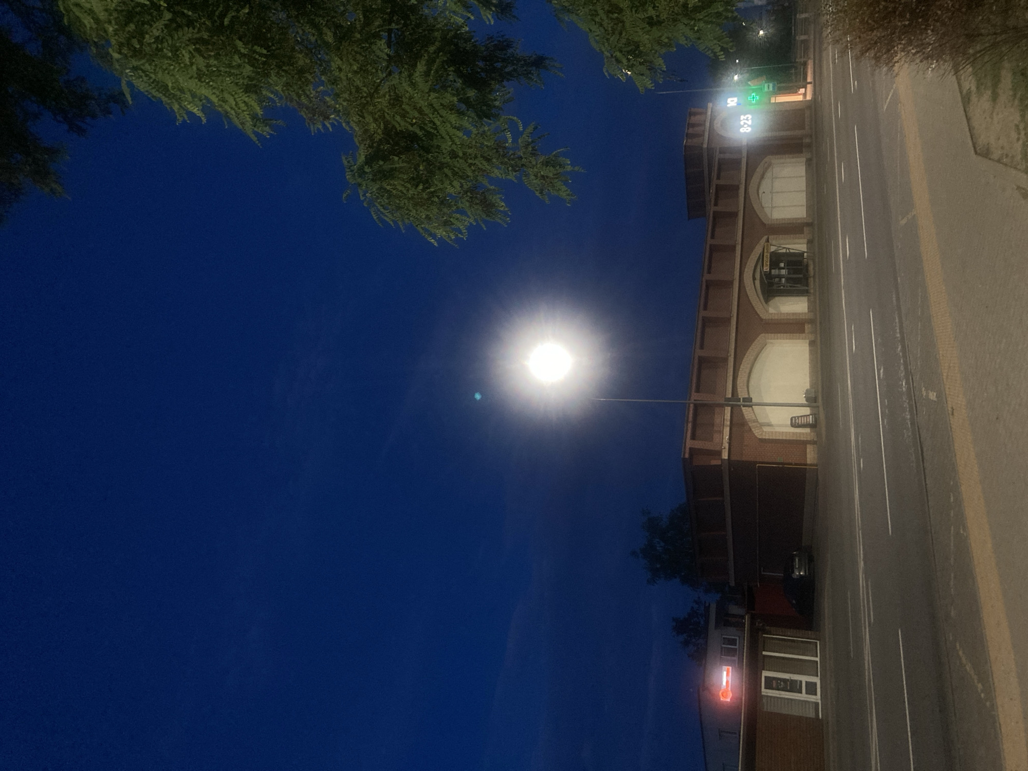 This image depicts a street scene at dusk or night in the city of Brest, Belarus. The sky is a deep, dark blue with some faint clouds. A bright street lamp dominates the upper center of the image, its light creating a halo effect. To the right, a large tree with dark green foliage hangs into the frame from the top.
In the middle ground, a low-rise building with a brick facade and arched window openings stretches across the frame. The windows appear to be either boarded up or covered. To the left of this building, a smaller, lighter-colored structure with a prominent neon sign reading "8-23" is visible, along with a green illuminated cross, suggesting a pharmacy. A dark-colored car is parked in front of the brick building.
The foreground shows a paved sidewalk and a road with white lane markings. The overall atmosphere is quiet and somewhat desolate, characteristic of a commercial area after business hours. There are no people visible in the image. The building with the "8-23" sign is likely a business, potentially a shop or a pharmacy, indicating its operating hours.