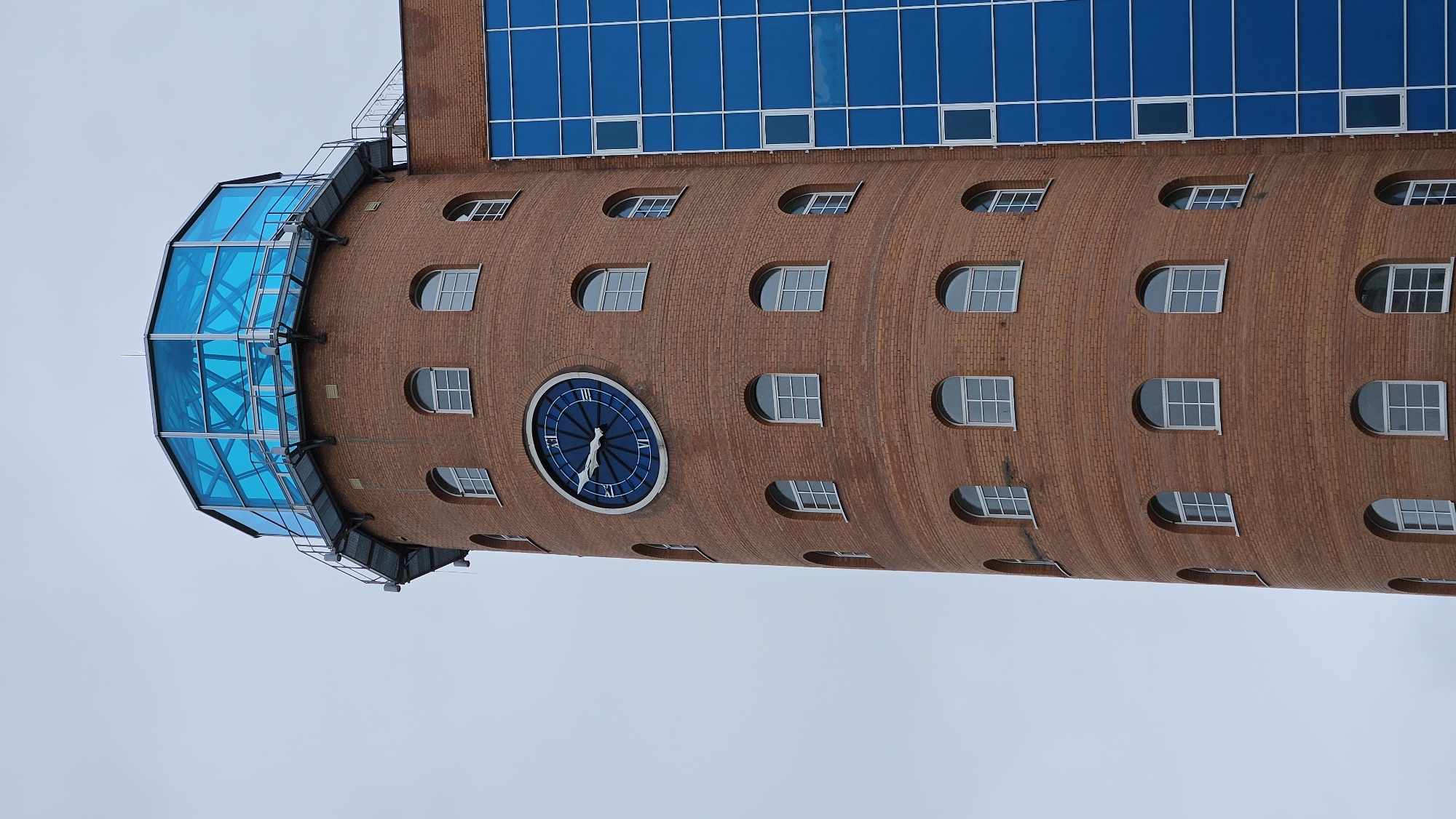 The image shows a close-up, low-angle view of a tall, cylindrical brick tower with a glass-enclosed observation deck at the top. The tower has numerous arched windows with white frames. A large, blue clock with Roman numerals is prominently displayed on the brick facade. Adjacent to the brick tower is a section of a modern building with a facade of blue glass panes. The sky is overcast and a uniform light gray.