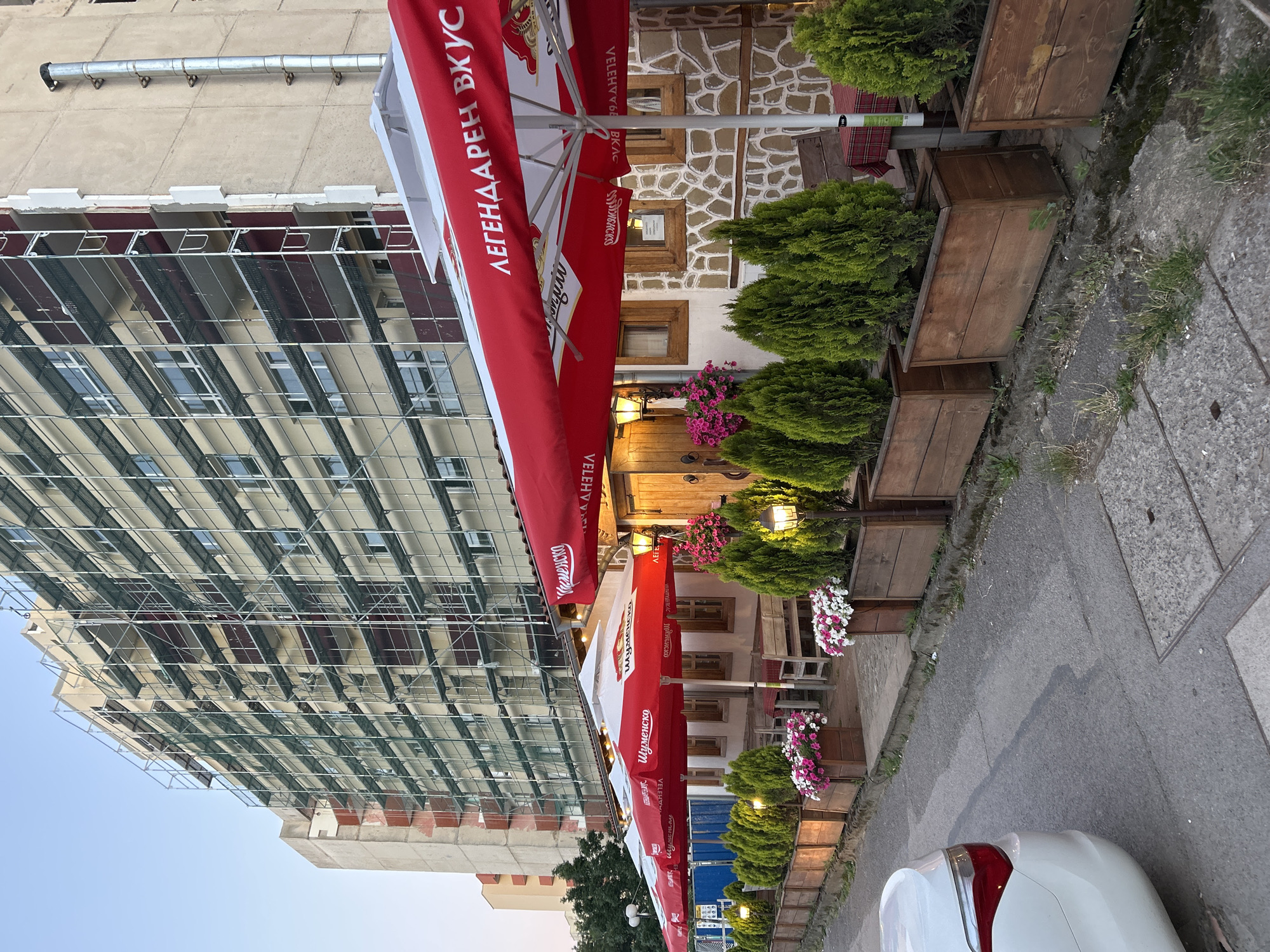 The image depicts an exterior view of a restaurant in Sofia, Bulgaria, with a building under renovation in the background. The foreground shows a sidewalk adjacent to the restaurant's outdoor seating area. Several red and white umbrellas, some bearing the text "Шуменско" and "ЛЕГЕНДАРЕН ВКУС", are set up over tables and chairs. Wooden planters filled with small, green coniferous trees and flowering plants line the sidewalk in front of the restaurant. The building behind the restaurant is covered in scaffolding, indicating ongoing construction or repair work. A white car is partially visible on the left side of the image. The overall scene suggests an urban setting, likely in the evening, given the lighting.
