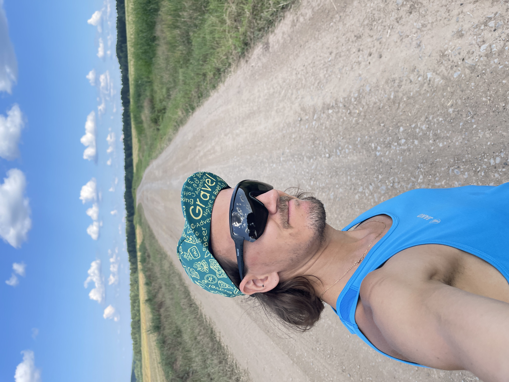 A man in a blue athletic tank top and a green cap with the word "Gravel" printed on it is taking a selfie on a dirt road. The road stretches into the distance, lined with grassy fields and trees under a blue sky with white clouds. The man is wearing black sunglasses that reflect the sky and clouds. He has medium-length brown hair and a mustache. A thin gold chain is visible around his neck. The setting appears to be rural, likely outside of Perevoznoe, Russia, given the natural landscape.