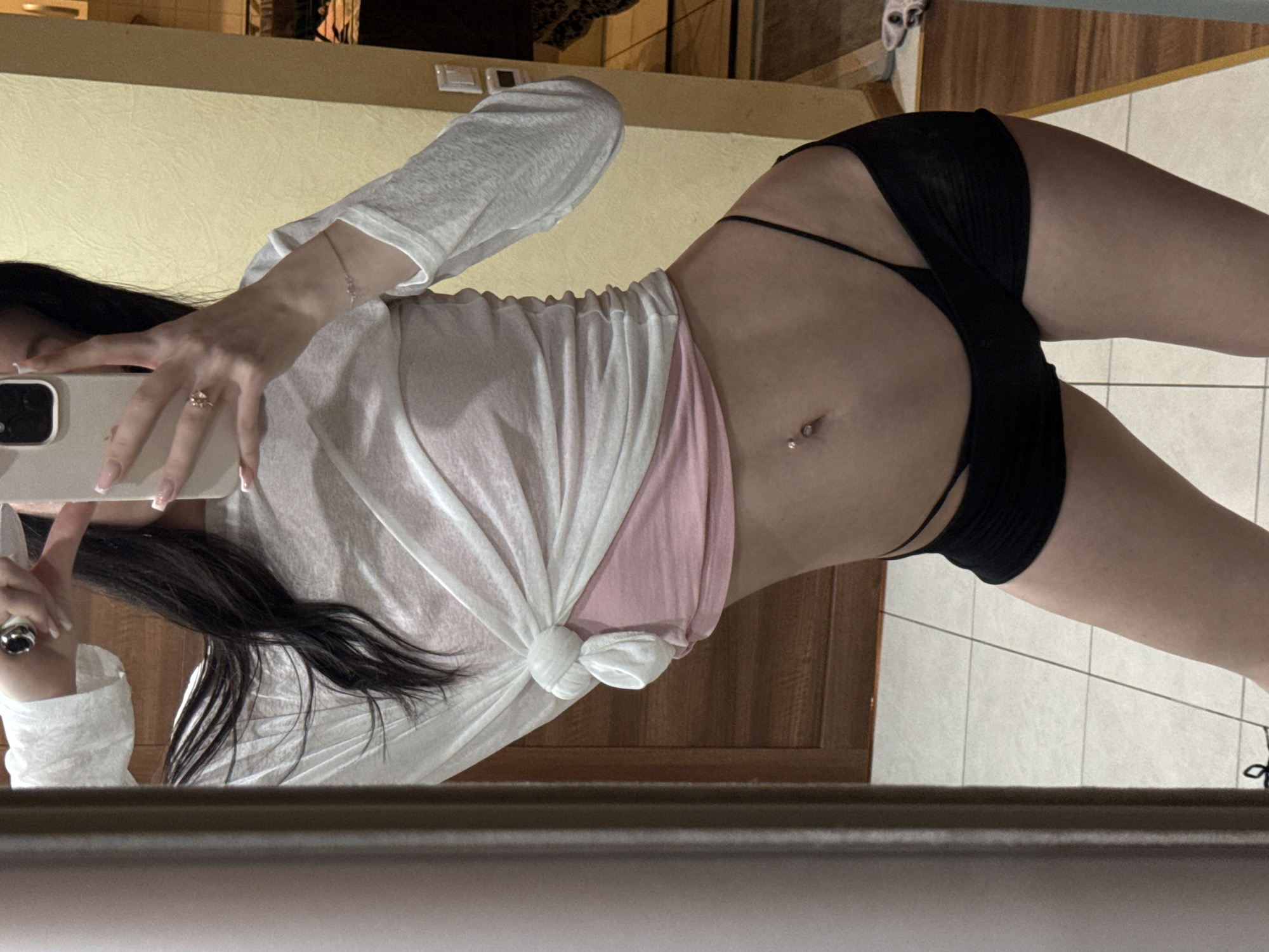 A woman is taking a mirror selfie in what appears to be a residential setting. She is wearing a white long-sleeved shirt tied at the waist, revealing a pink camisole underneath. She also has on black shorts and has a belly button piercing. The background shows a wooden door and a tiled floor, with a partial view of what looks like a hallway. There is a phone in her hand, and she is wearing rings and a bracelet.