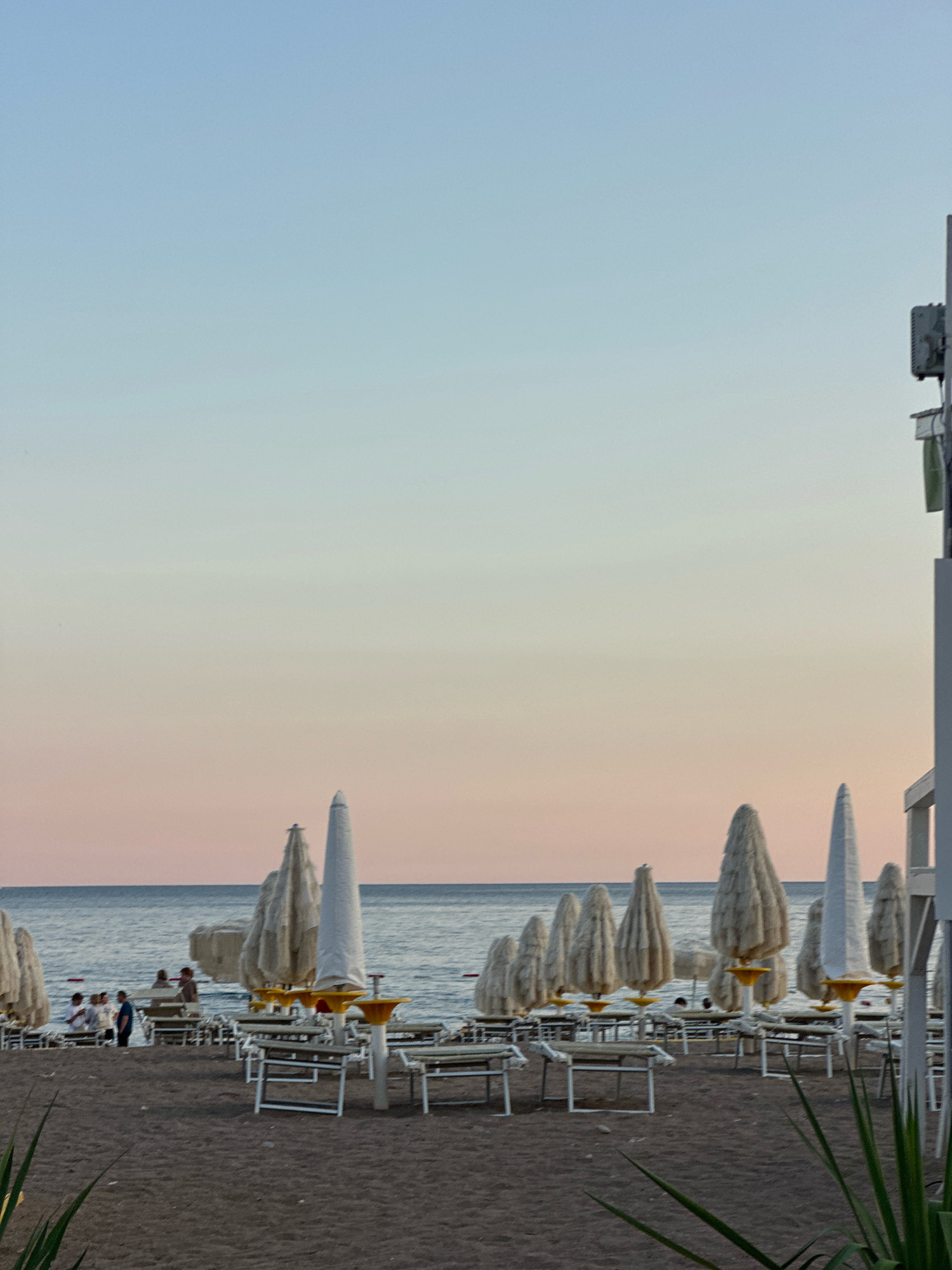 The image depicts a beach scene at sunset in Boreti, Montenegro. The sand is dark, and numerous beach chairs and umbrellas are set up. In the distance, the sea is visible, with a soft pastel-colored sky above. Several people are scattered across the beach, some sitting on chairs and others standing. The umbrellas are mostly closed and appear to be made of canvas. A tall structure, possibly a lifeguard stand or a part of a building, is visible on the right side of the frame. The overall atmosphere is serene, with the warm light of the setting sun casting a gentle glow on the scene.
