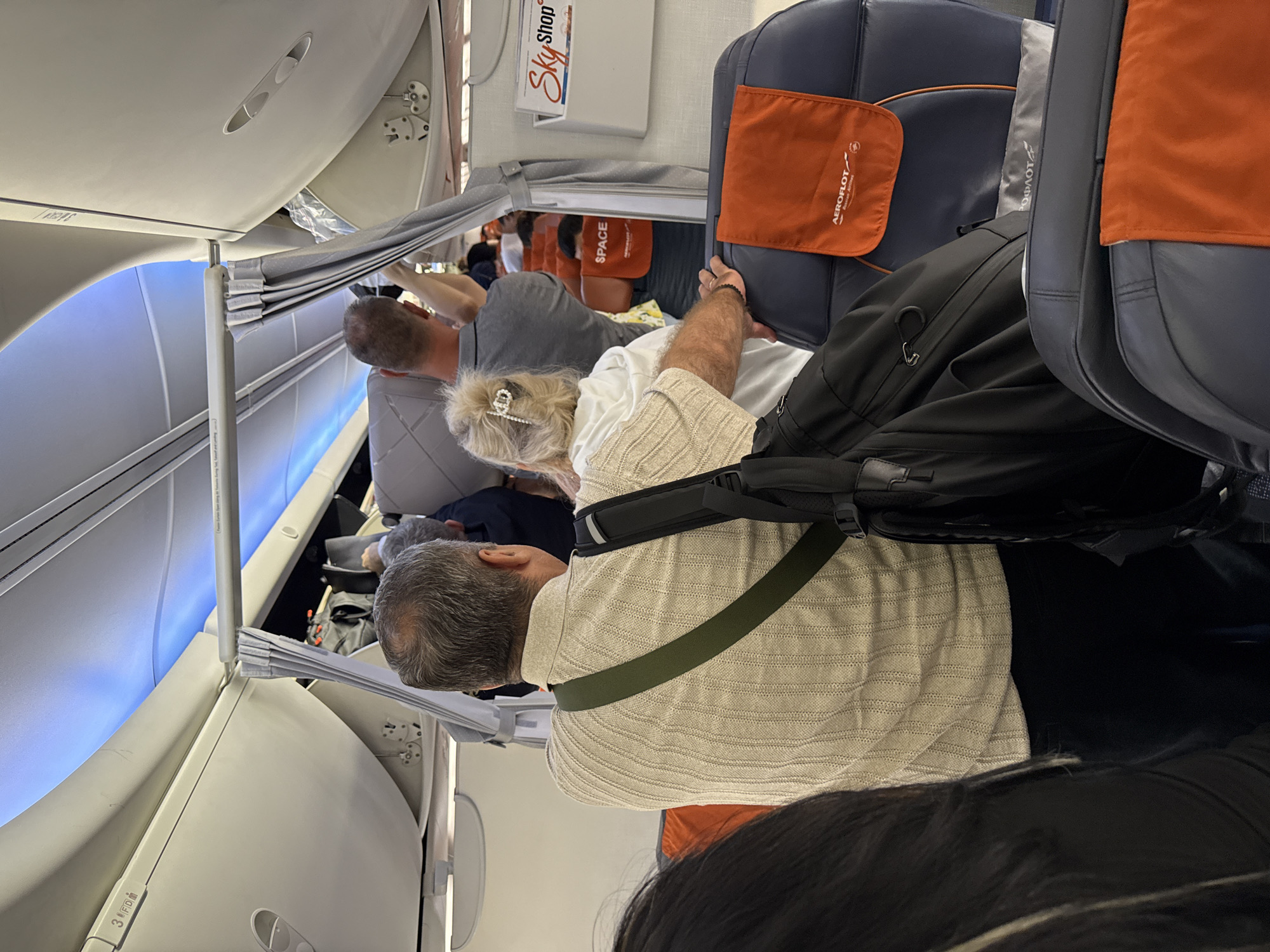 An interior view of a crowded airplane aisle shows passengers moving about. In the foreground, a man with gray hair and wearing a beige sweater is seen from the back, with a green strap across his shoulder and a black backpack. To his right, a woman with blonde hair pulled back with a sparkly clip is wearing a white hoodie and is facing away from the camera. Behind them, a man in a gray shirt is visible, and further down the aisle, other passengers are seen among the seats. The overhead luggage bins are open, with some items being stored. A "Sky Shop" magazine is visible on the wall to the right. The seats are upholstered in dark blue with orange headrests, some of which have the Aeroflot logo. The lighting suggests an evening flight, with blue ambient lights along the ceiling. The image appears to have been taken on a Boeing 737 aircraft at Istanbul Airport, located in Arnavutköy, Turkey, likely during boarding or disembarking procedures.