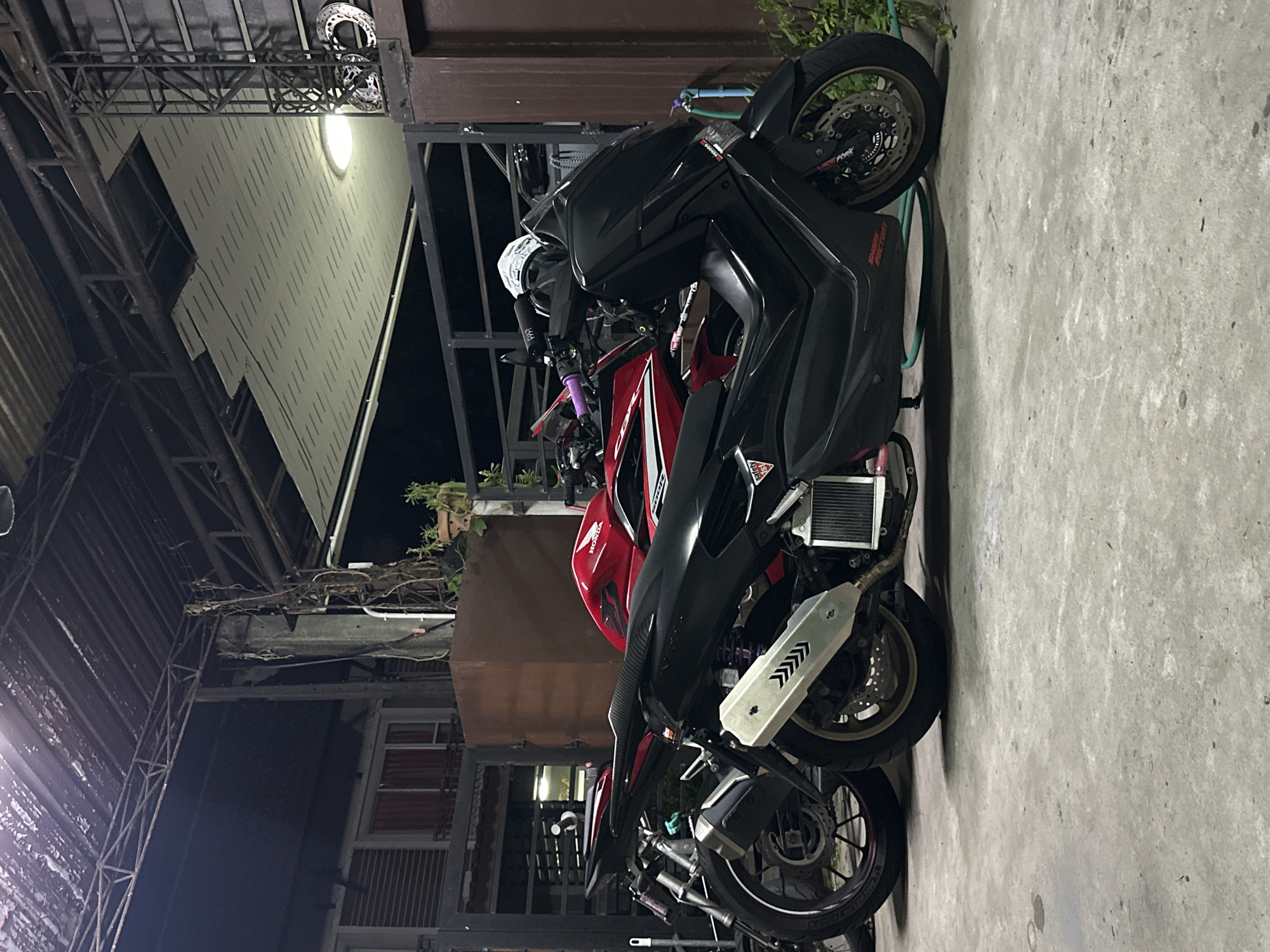 This media file depicts a still scene of two motorcycles parked next to each other. The motorcycles are the primary subjects, with one being a larger, black sportbike and the other a red and black sportbike. They appear to be parked under a covered structure, possibly a carport or the overhang of a building, with a concrete ground surface.
The setting is outdoors at night, as indicated by the artificial lighting and the dark sky. The lighting casts shadows and highlights on the motorcycles and the surrounding environment. The scene appears to be in Kathu, Thailand, suggesting a warm climate. There are hints of vegetation in the background.
Notable details include the intricate design of the motorcycles' wheels and exhausts, and the presence of what appears to be a water hose coiled on the ground near the red motorcycle. Some text is visible on the motorcycles, including "HONDA" on the red one, and other decals on the black sportbike that are partially obscured. There is a circular disc, possibly a brake rotor, mounted high on a metal structure in the background, near a light source.
No people are visible in the image. The overall mood is quiet and still, capturing a moment of repose for the vehicles. The scene is likely a residential or commercial area where motorcycles are commonly used and stored.