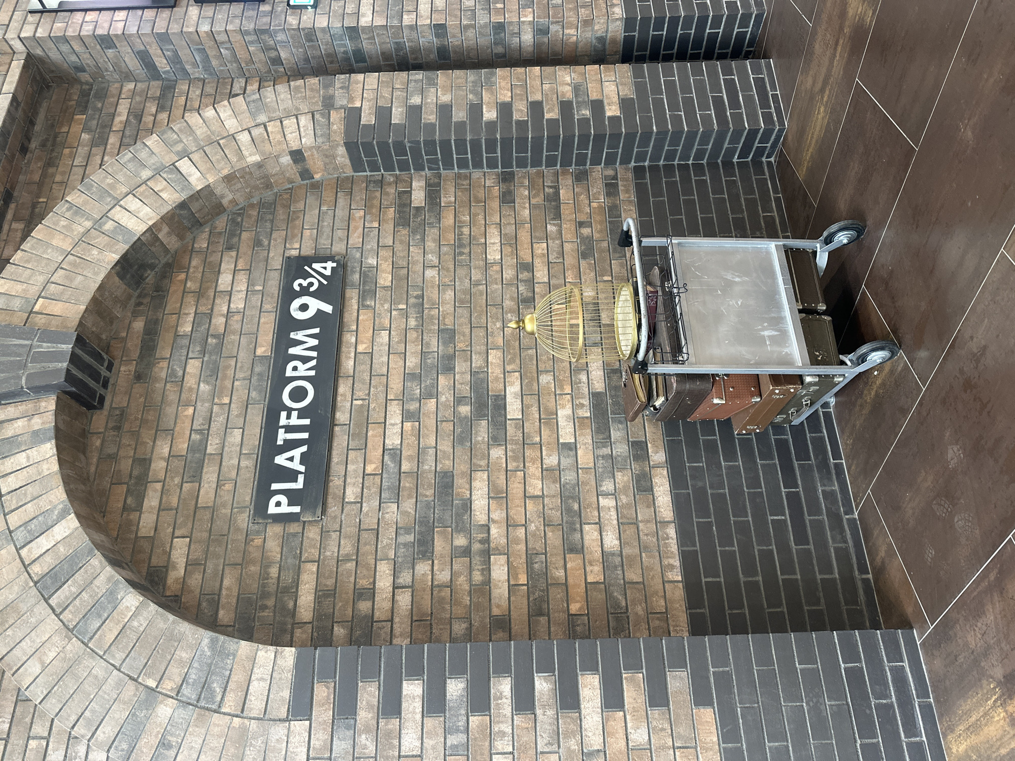 A cart laden with vintage suitcases and a golden birdcage sits on a tiled floor in front of a brick wall. Above the cart, a sign reads "PLATFORM 9 3/4" in white lettering on a black background. The wall features an arched brick structure. The scene appears to be a replica of the iconic Harry Potter Platform 9 ¾ at King's Cross Station in London.
