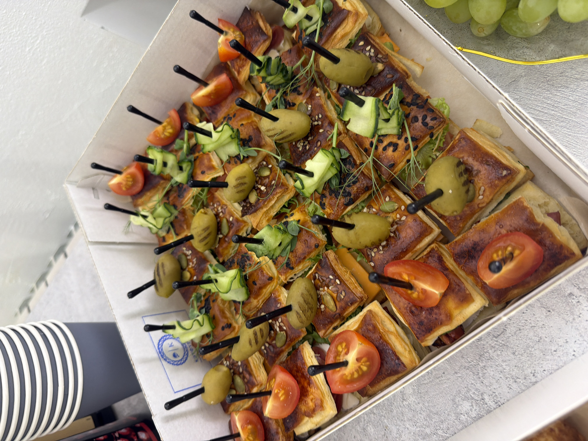 The image shows a close-up of a box filled with square-shaped appetizers, likely served during an event or gathering. The appetizers are topped with various ingredients, including sliced tomatoes, olives, and cucumber ribbons. Black skewers are inserted into each appetizer, presumably for easy handling. Some of the appetizers appear to have a pastry or bread base. There is a container of grapes partially visible in the upper right corner. The scene is well-lit, suggesting an indoor setting. The location in Chaikovsky, Russia, is not discernible from the image.