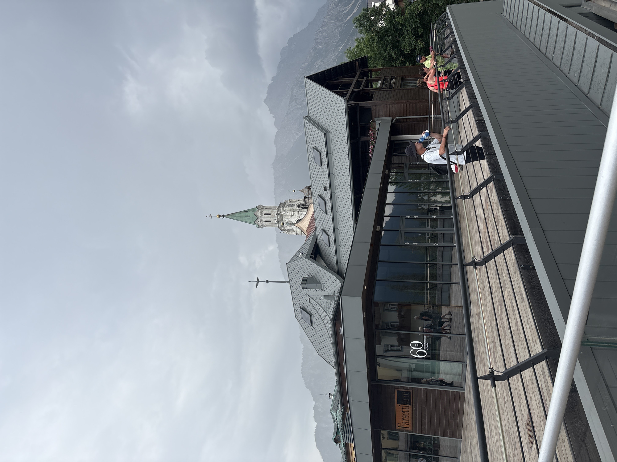 The image shows a modern building with a wooden deck and glass facade, set against a backdrop of mountains and a cloudy sky. Several people are visible on the deck, some appearing to be taking a break or observing the scenery. One person is leaning on a railing and drinking from a water bottle. Another person is wearing a bright orange top, and others are in casual clothing. The building has multiple levels and a distinctive grey, patterned roof. A church spire with a green roof is visible in the distance, suggesting a town or city setting nestled near the mountains. The weather appears overcast, and the overall atmosphere is relaxed. Text visible on the building's facade includes "60" and "Farsetti". The image is oriented vertically.