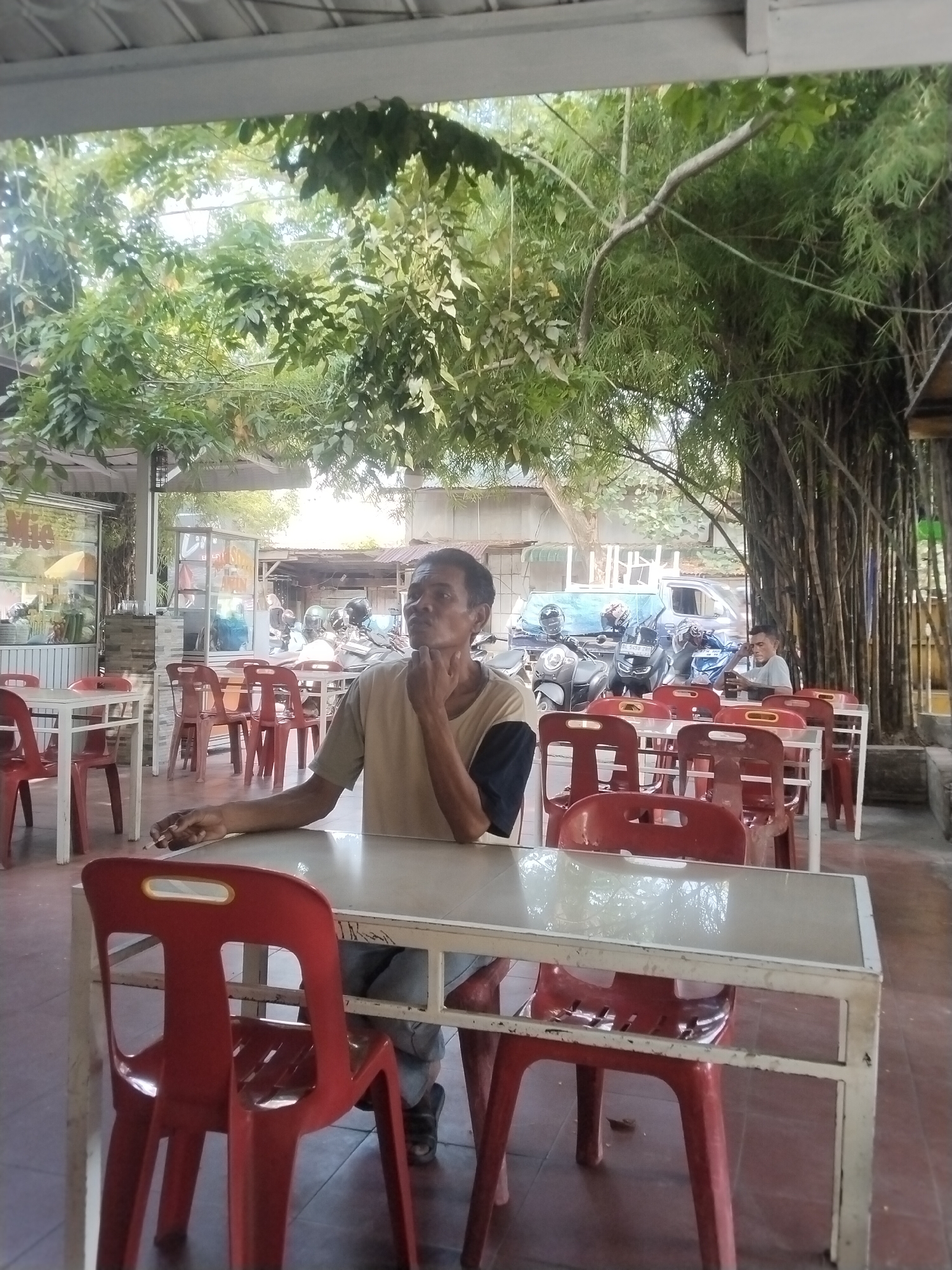 A man sits at a white, rectangular table with red plastic chairs in an open-air restaurant in Lhokseumawe, Indonesia. He wears a light brown t-shirt with a dark blue sleeve. His left hand is at his chin, and his right hand holds a lit cigarette on the table. Other red chairs and white tables populate the restaurant. In the background are motorbikes, parked vehicles, and another person sitting at a table. Bamboo trees are visible at the far right. Vegetation grows overhead. The ground is red tile.