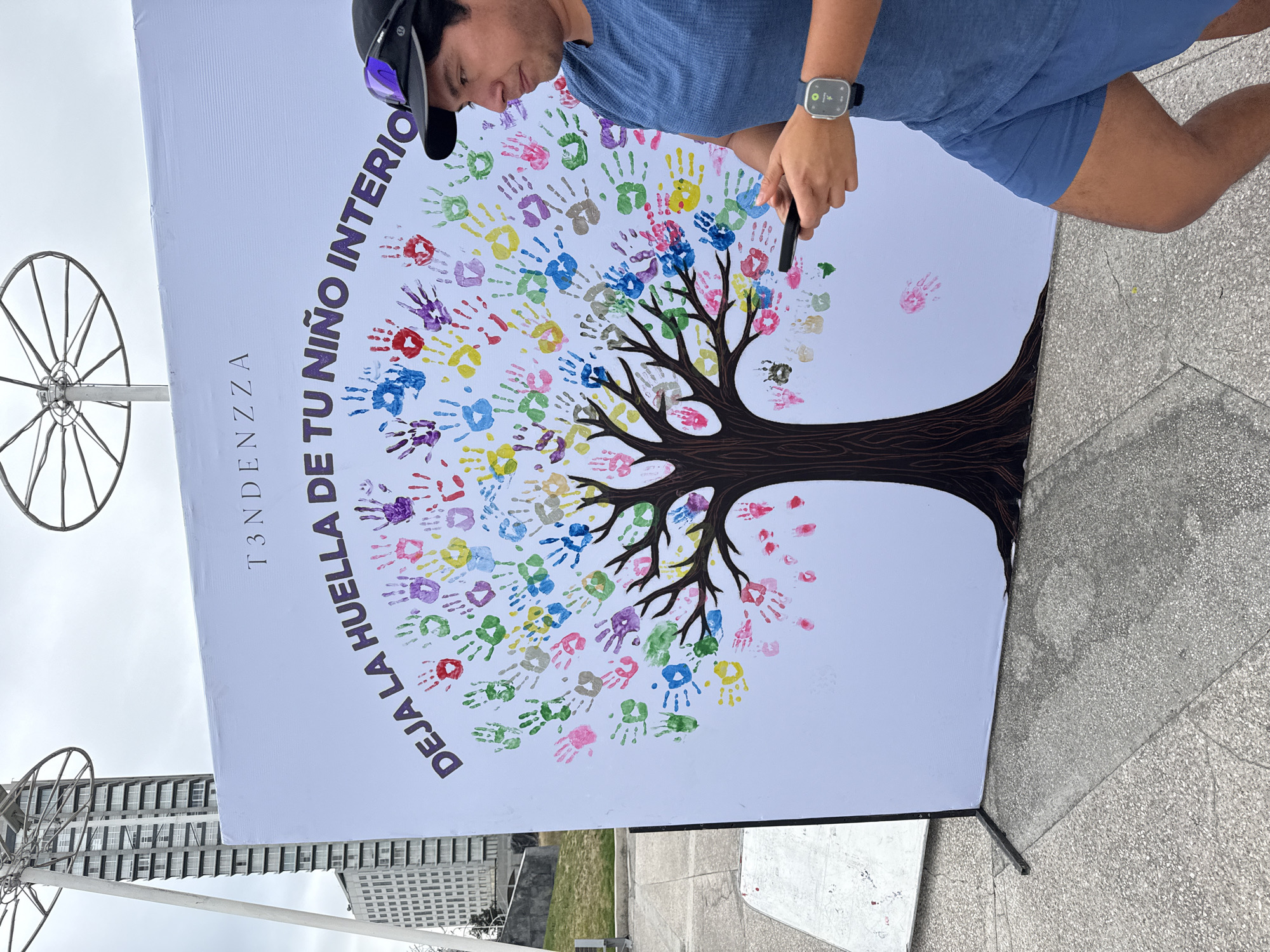 A man stands to the right of a poster displaying a tree with colorful handprints for leaves. The poster reads "DEJA LA HUELLA DE TU NIÑO INTERIOR" and "T3NDENZZA". The man is wearing a blue shirt, blue shorts, a black cap, and a smartwatch. He is holding a black object. The poster is positioned on a sidewalk in what appears to be an urban setting. In the background, there is a building and a decorative wheel structure. The location is Puebla, Mexico.