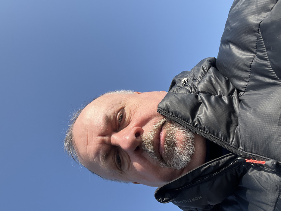 A low-angle, head-and-shoulders portrait depicts an adult male against a clear, bright blue sky, indicating an outdoor, daytime setting in the city of Troitsk, Russia. The man has fair skin, graying hair visible on the crown, and a graying goatee-style beard. His eyes are open, directed forward, and his facial expression is neutral. He is wearing a dark, possibly black or dark gray, quilted winter jacket with a high collar, partially unzipped. The brand name "NORTHLAND" is visible vertically on the left side of the jacket's collar. A red toggle is attached to the main zipper pull. No other individuals, objects, or environmental features are visible to determine a specific location within Troitsk.