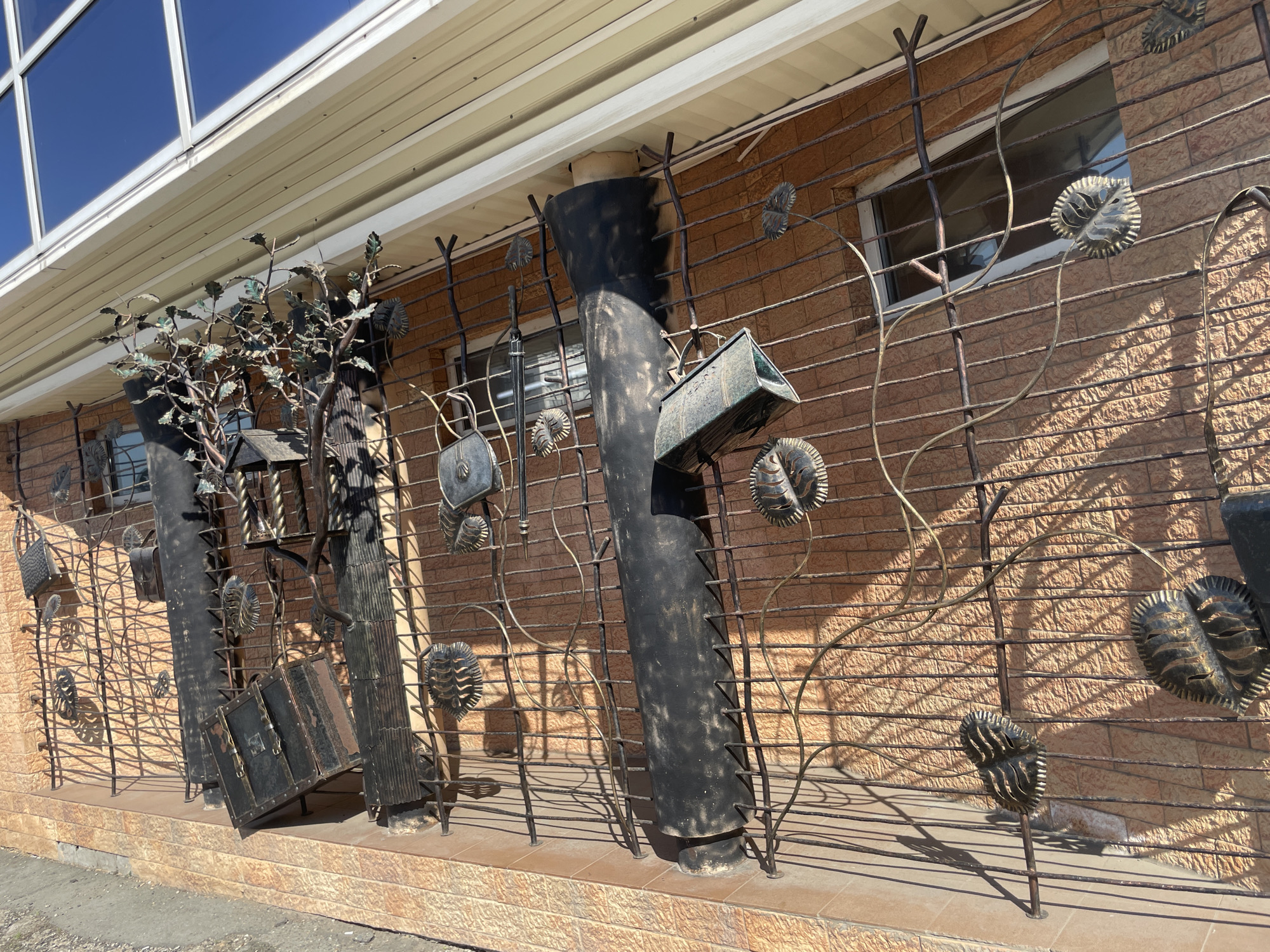 The image shows a decorative metal fence in front of a brick building. The fence is made of thin metal rods and features decorative elements such as leaves and stylized objects. The location is Izhevsk, Russia.